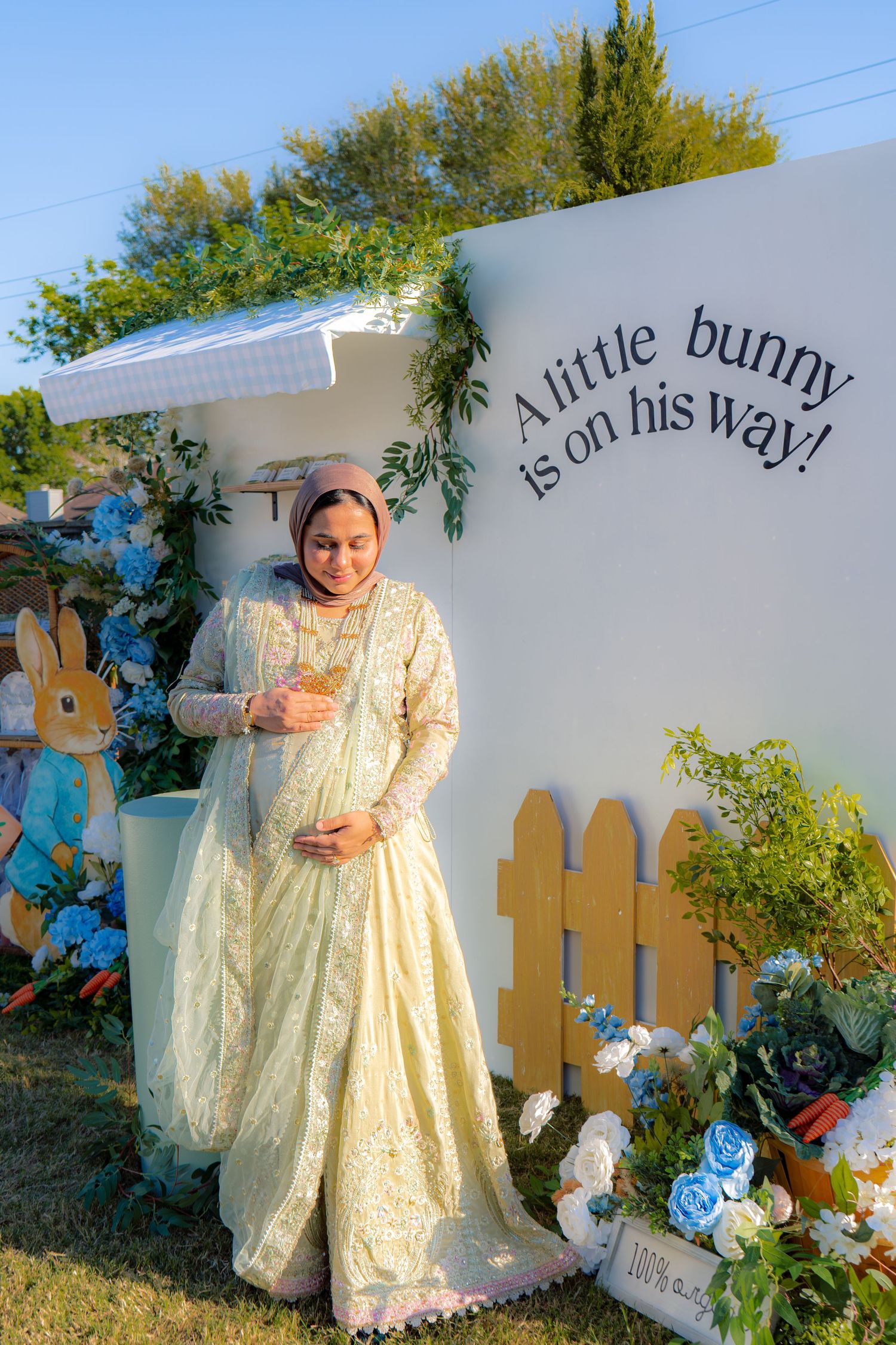 Pregnant woman in cream traditional dress posing at Peter Rabbit baby shower backdrop outdoors.