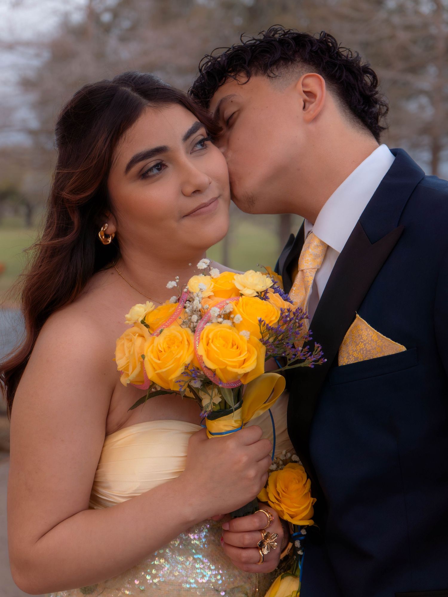 Prom couple sharing romantic moment with young man kissing girls cheek holding yellow roses.