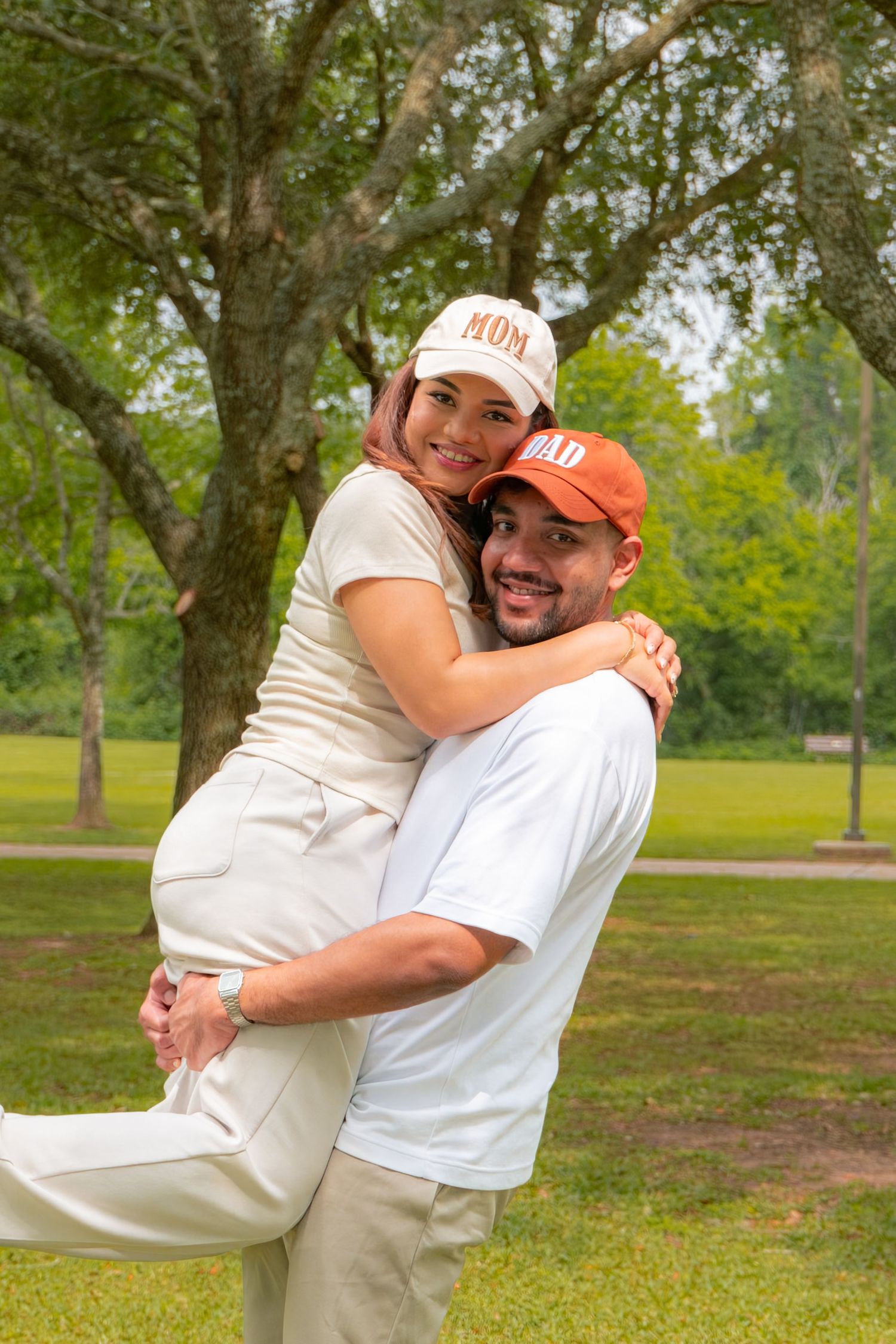 Excited couple wearing Mom and Dad caps, man lifting woman in a joyful embrace in a green park.