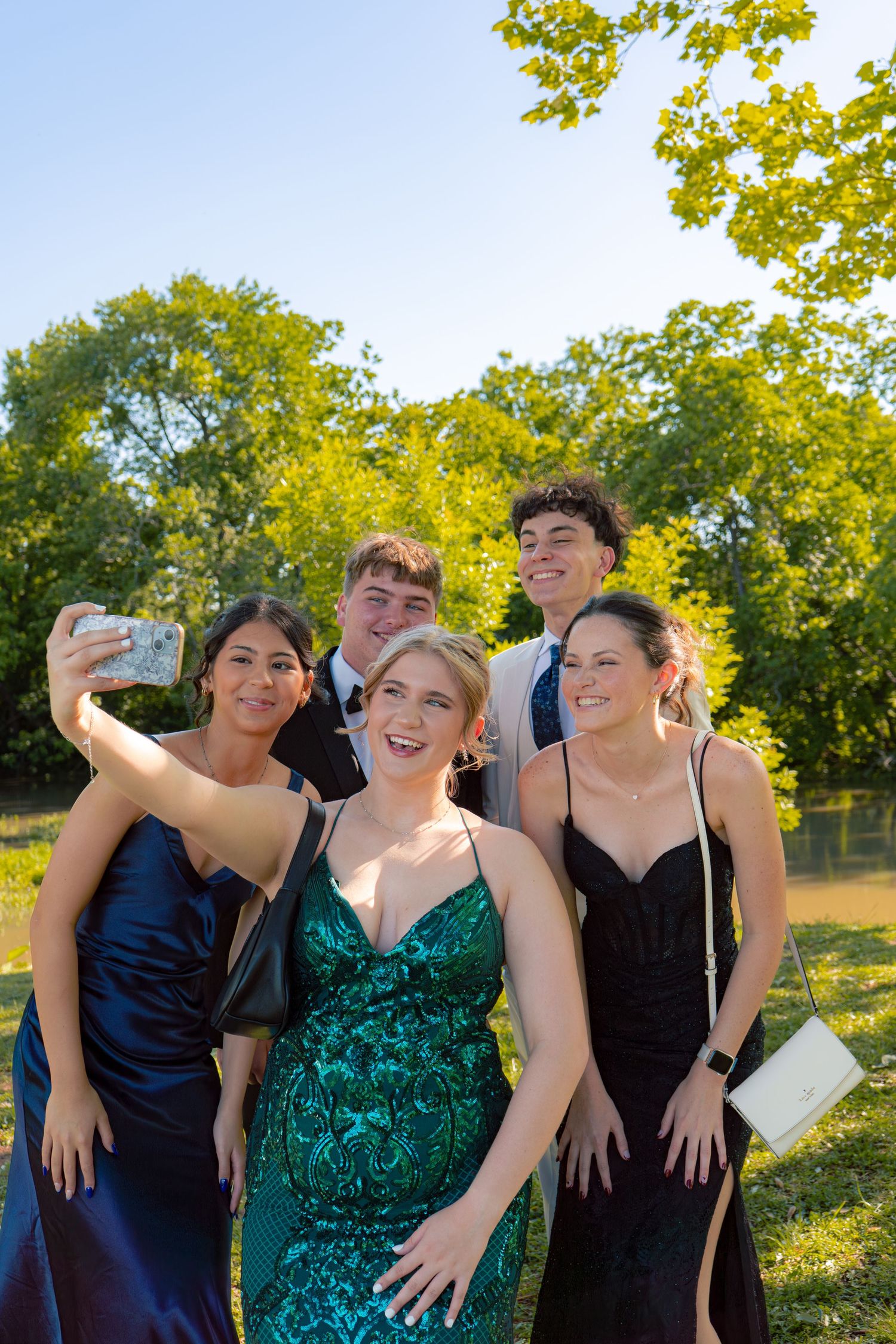 Group of five teenagers in prom attire take a selfie outdoors under bright sunny tree canopy.
