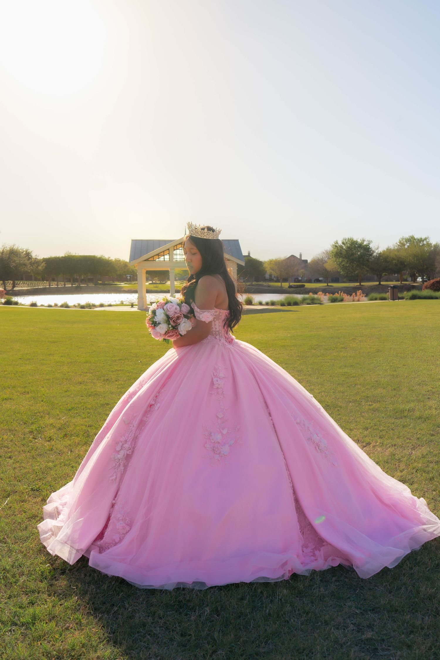 Young woman in a pink ball gown quinceañera dress holding a bouquet in a sunny park setting.