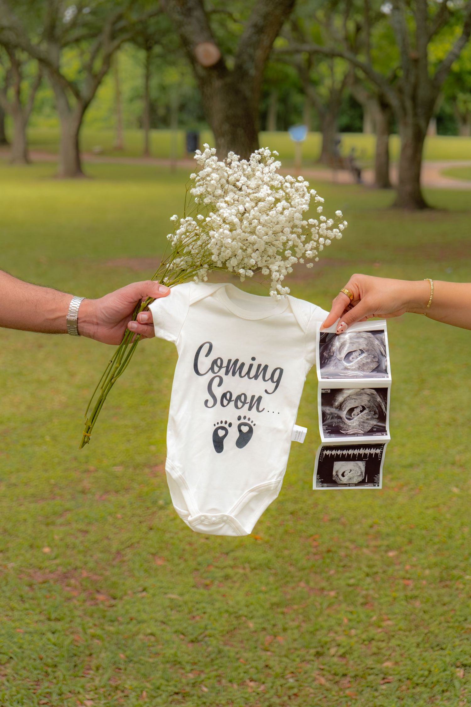 Couple holding baby onesie saying Coming Soon with ultrasound photos and white flowers in a park for pregnancy announcement.