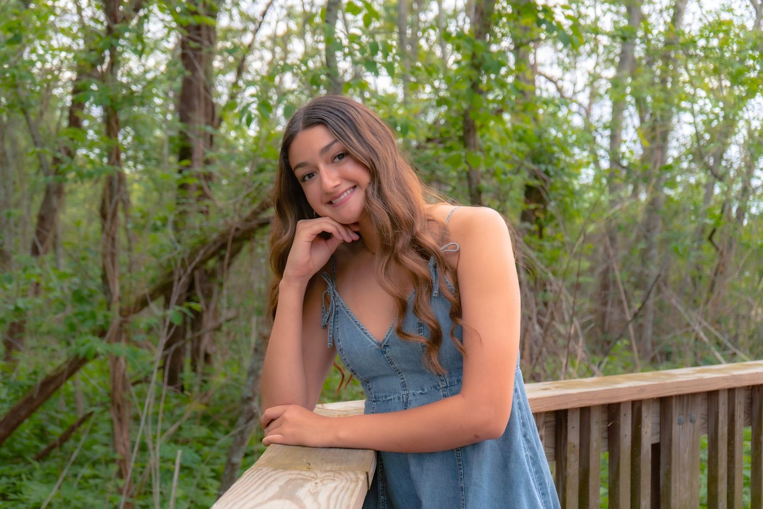 Young woman in denim dress smiling on wooden deck surrounded by lush green trees in outdoor nature setting.