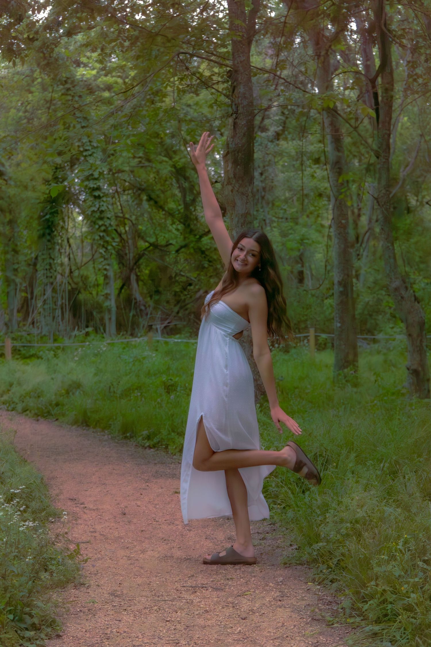 Young woman in a white maxi dress dancing joyfully on a forest trail with one arm raised.
