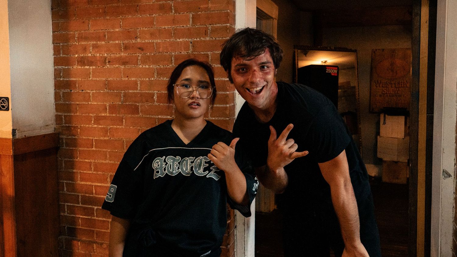Two people smiling and posing together in front of a brick wall indoors.