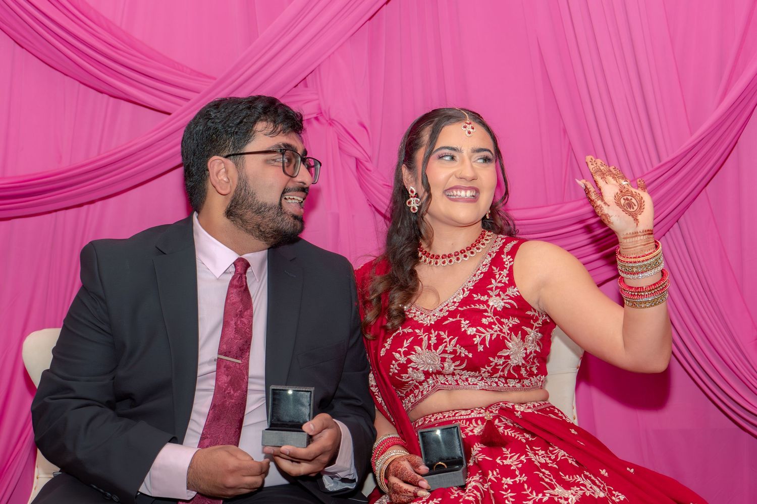 Couple in Indian wedding attire, woman in red lehenga with jewelry, man in dark suit, pink backdrop.