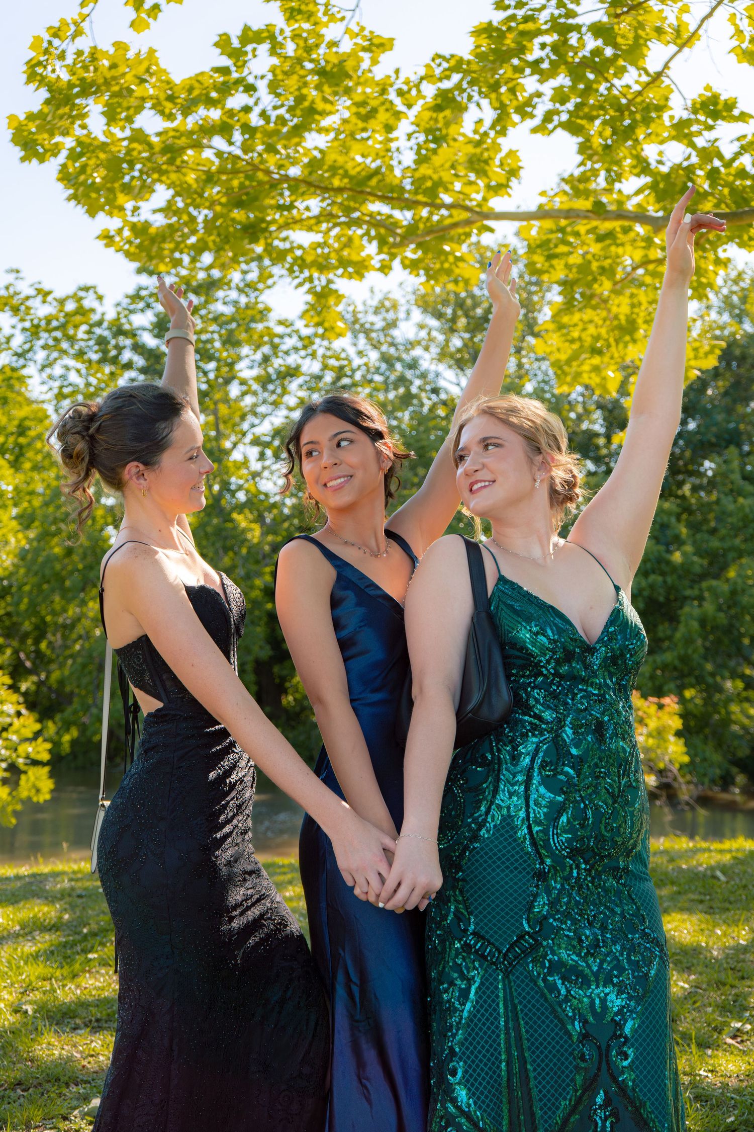 Three young women in black, navy, and green formal gowns hold hands and raise arms joyfully under tree branches.