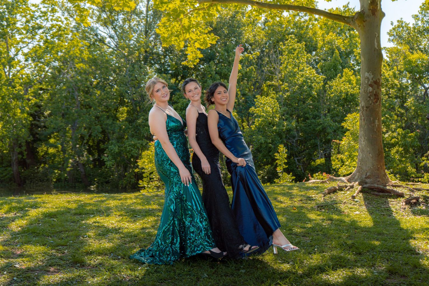 Three young women in elegant prom gowns pose outdoors in green and navy dresses among lush trees.