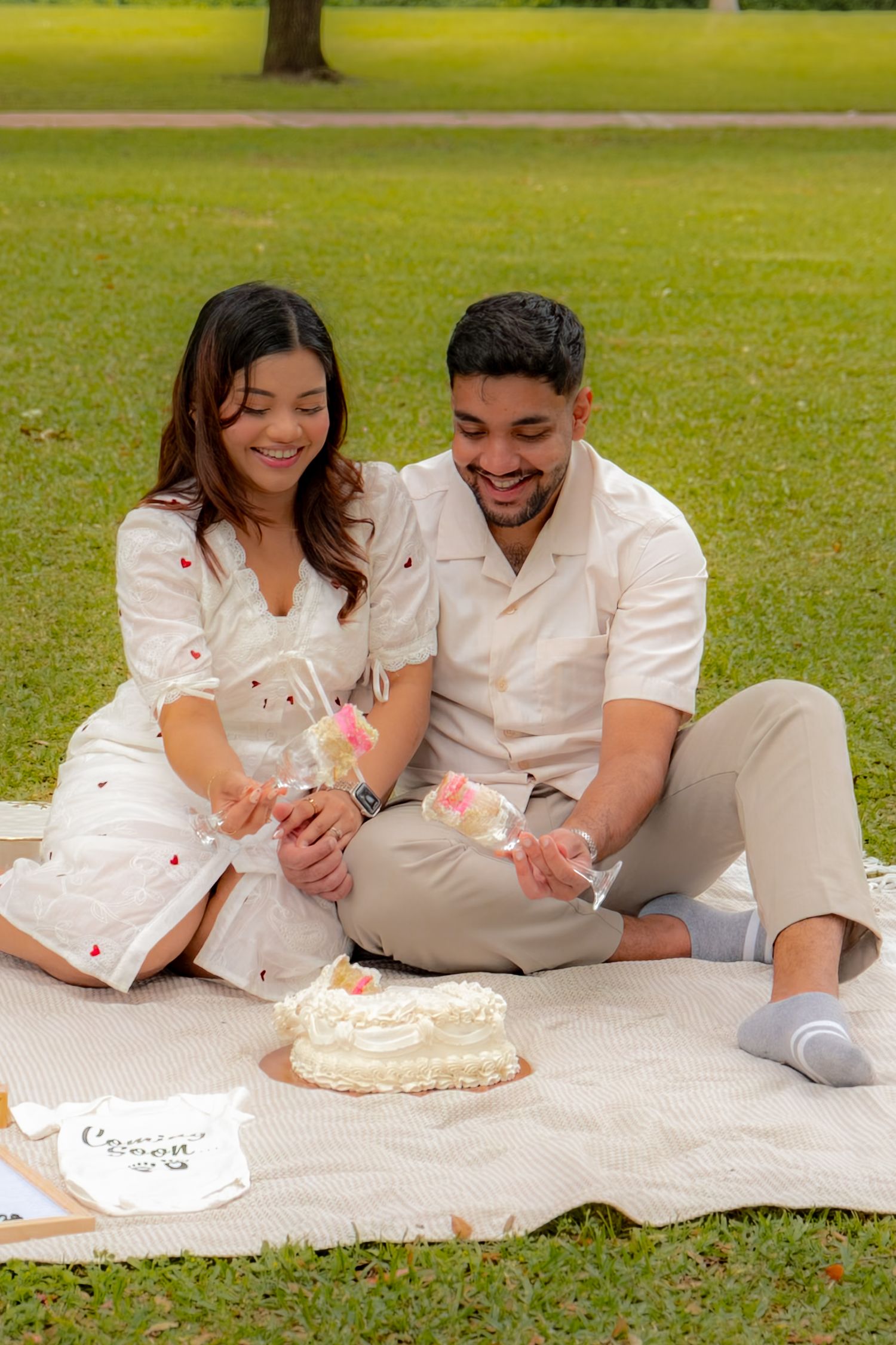 Happy couple sitting on picnic blanket in park holding cake toppers beside a white heart-shaped cake.