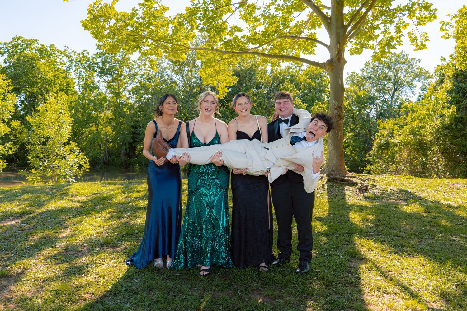 Five teens in formal attire pose outdoors, with four standing while lifting one person horizontally in a fun prom photo.