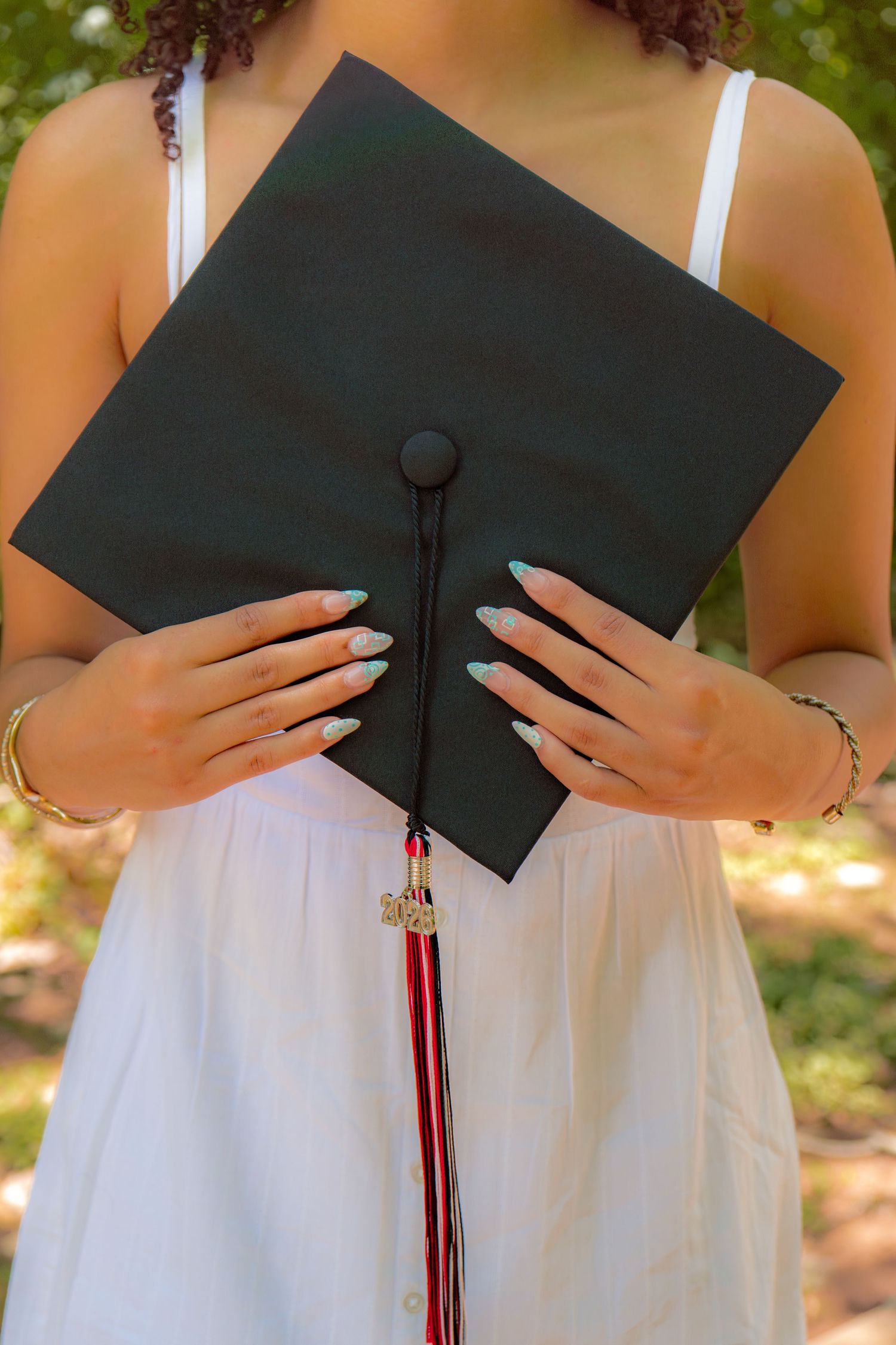 Young woman in white dress holding a black graduation cap with red and black tassel outdoors.