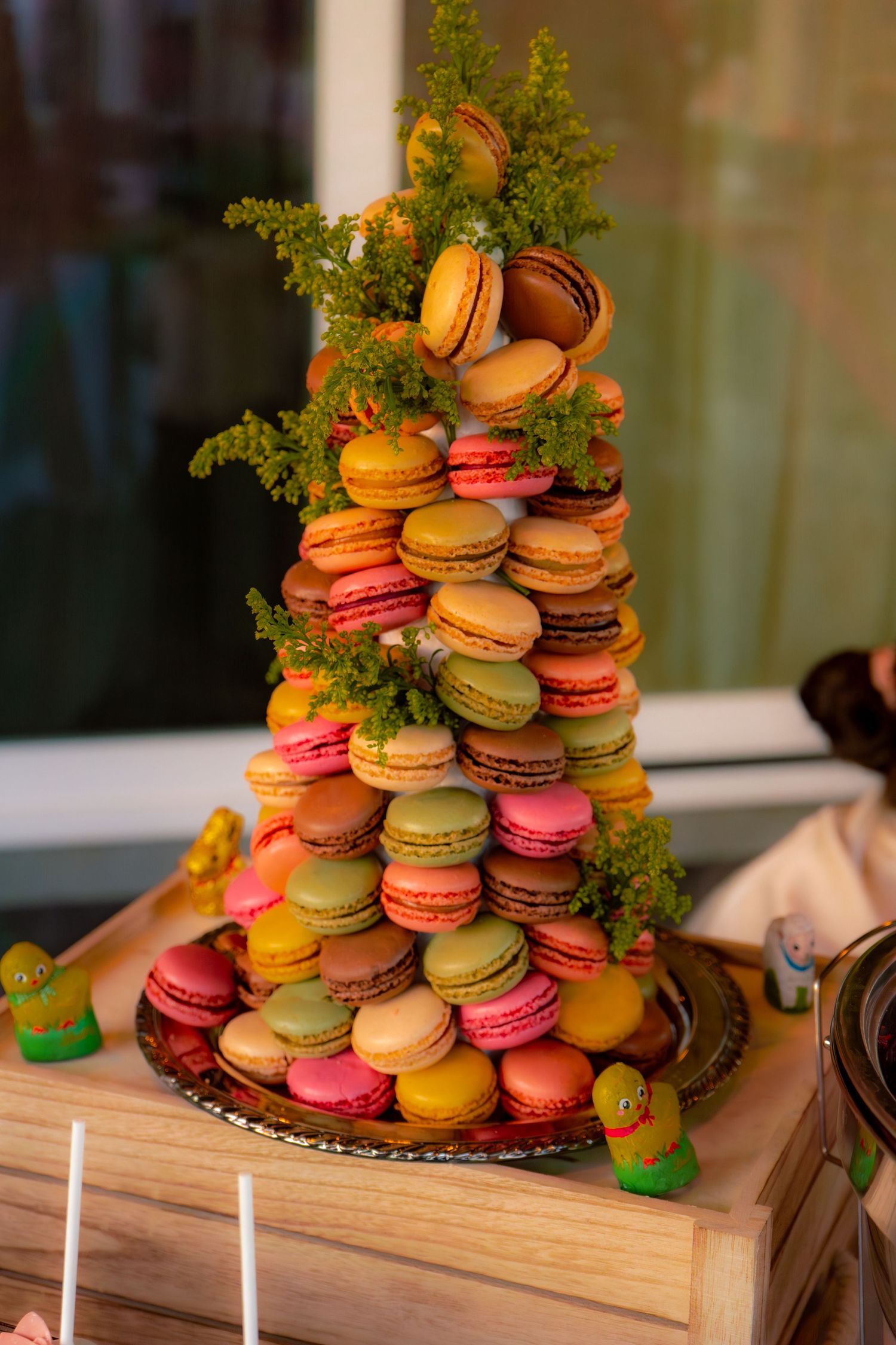 Colorful tower of French macarons in pink, orange, green, and brown stacked with green herb decorations.