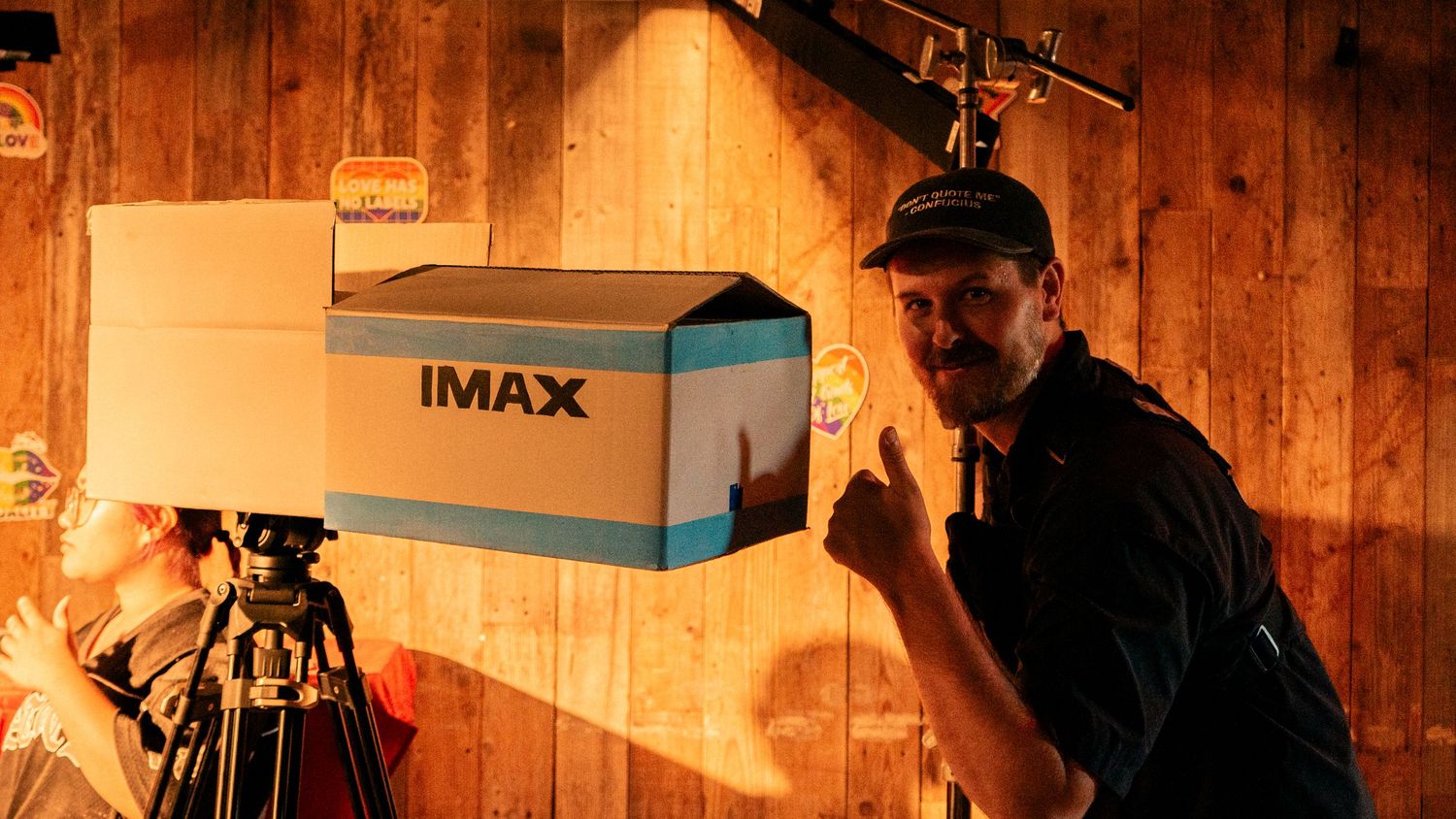 Man posing proudly next to an IMAX camera box on a film set with warm lighting and equipment.