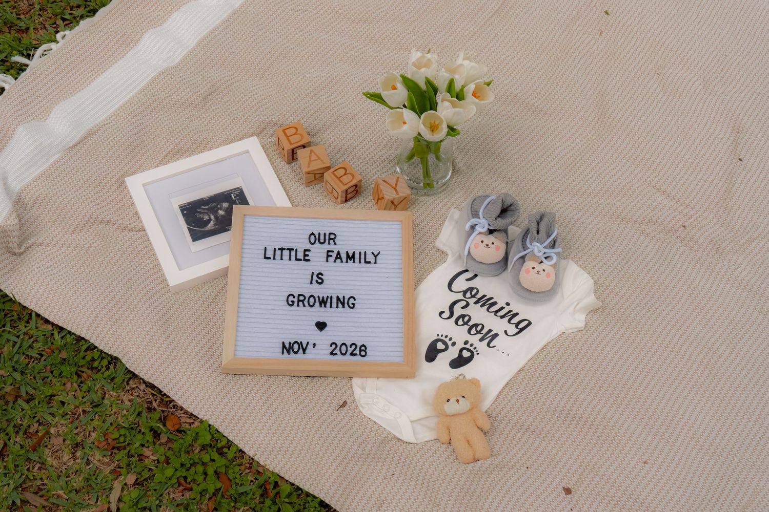 Pregnancy announcement flat lay with letter board reading Our Little Family is Growing Nov 2028, baby shoes, onesie, and flowers.