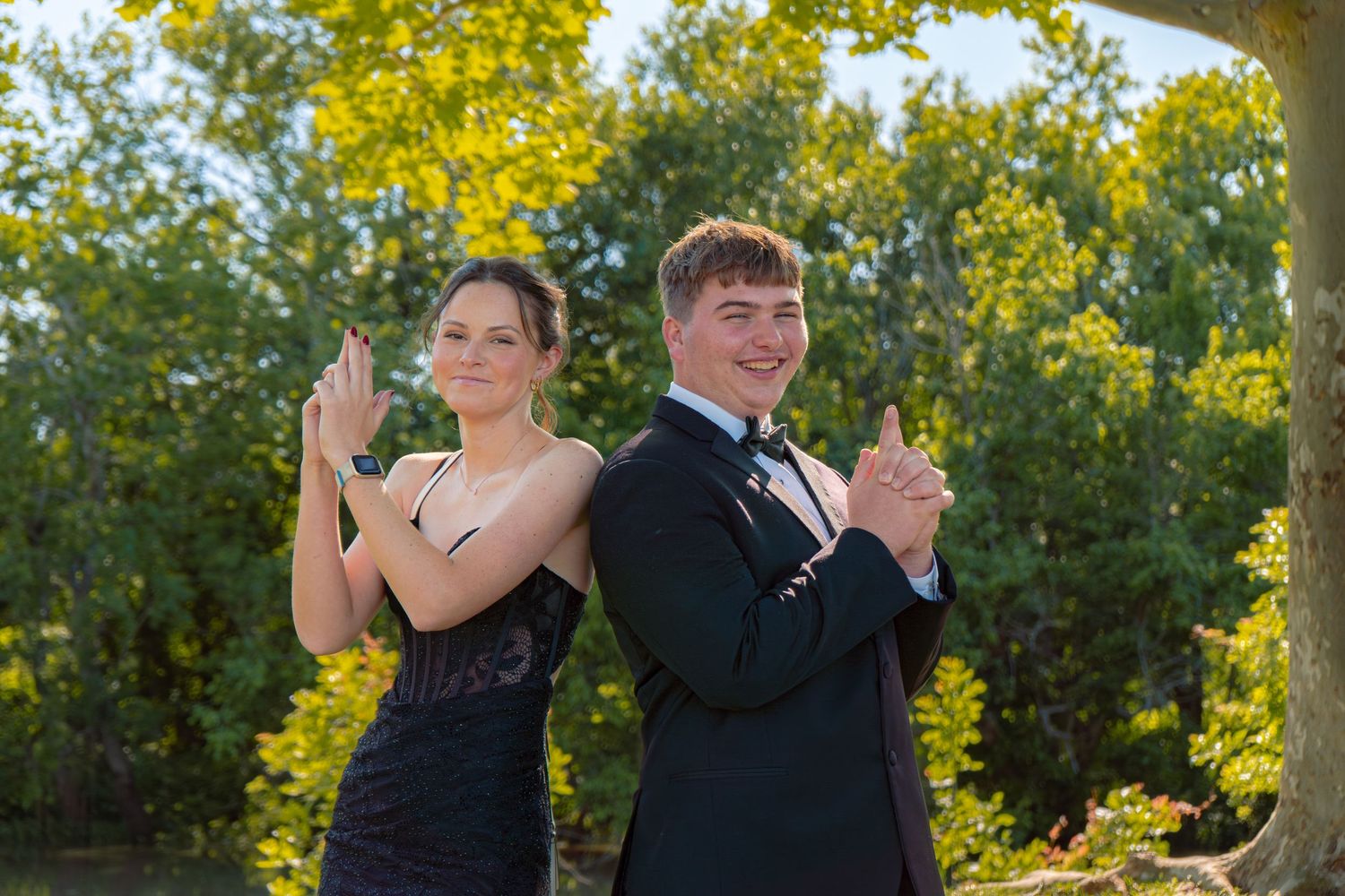 A young couple in formal prom attire poses back-to-back outdoors, both making stylish hand gestures with green trees behind them.