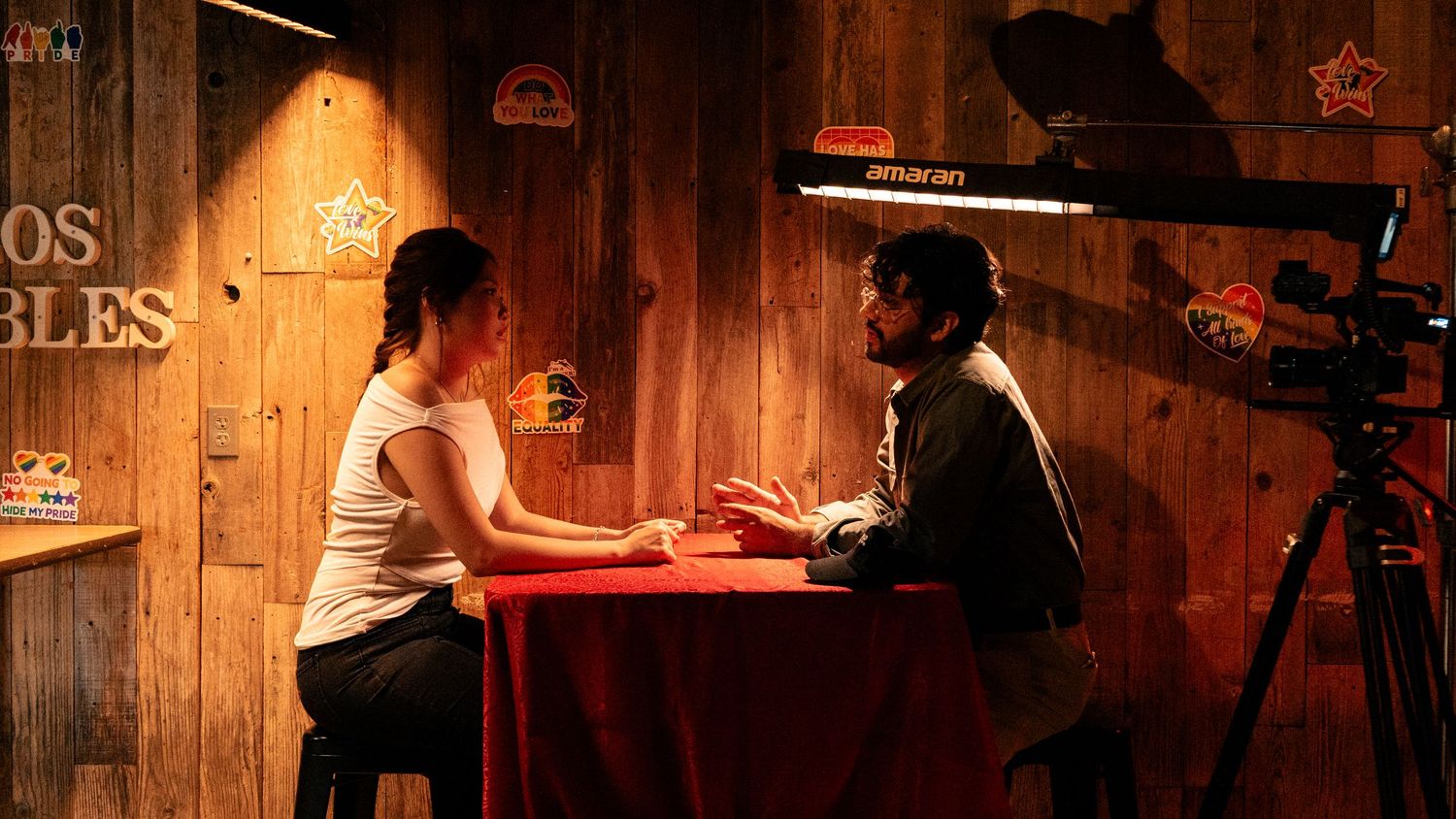 Two people sit at a red table in a warmly lit wooden studio during a filmed interview with camera on tripod.