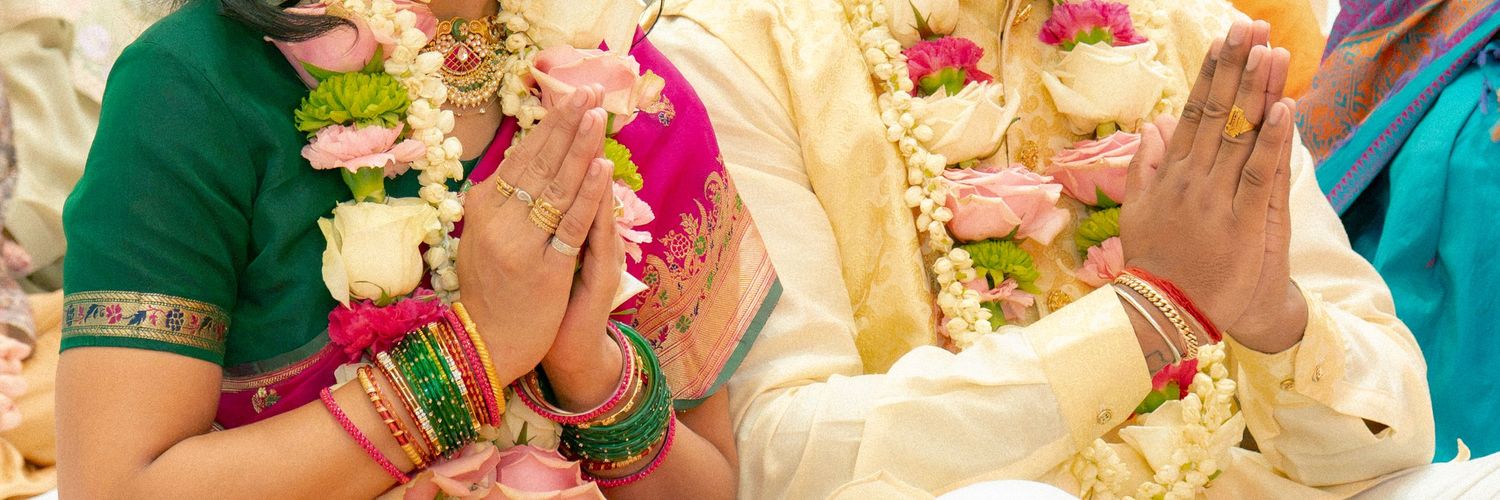 Indian wedding couple in traditional attire with floral garlands performing sacred ceremony rituals with hands raised in prayer.