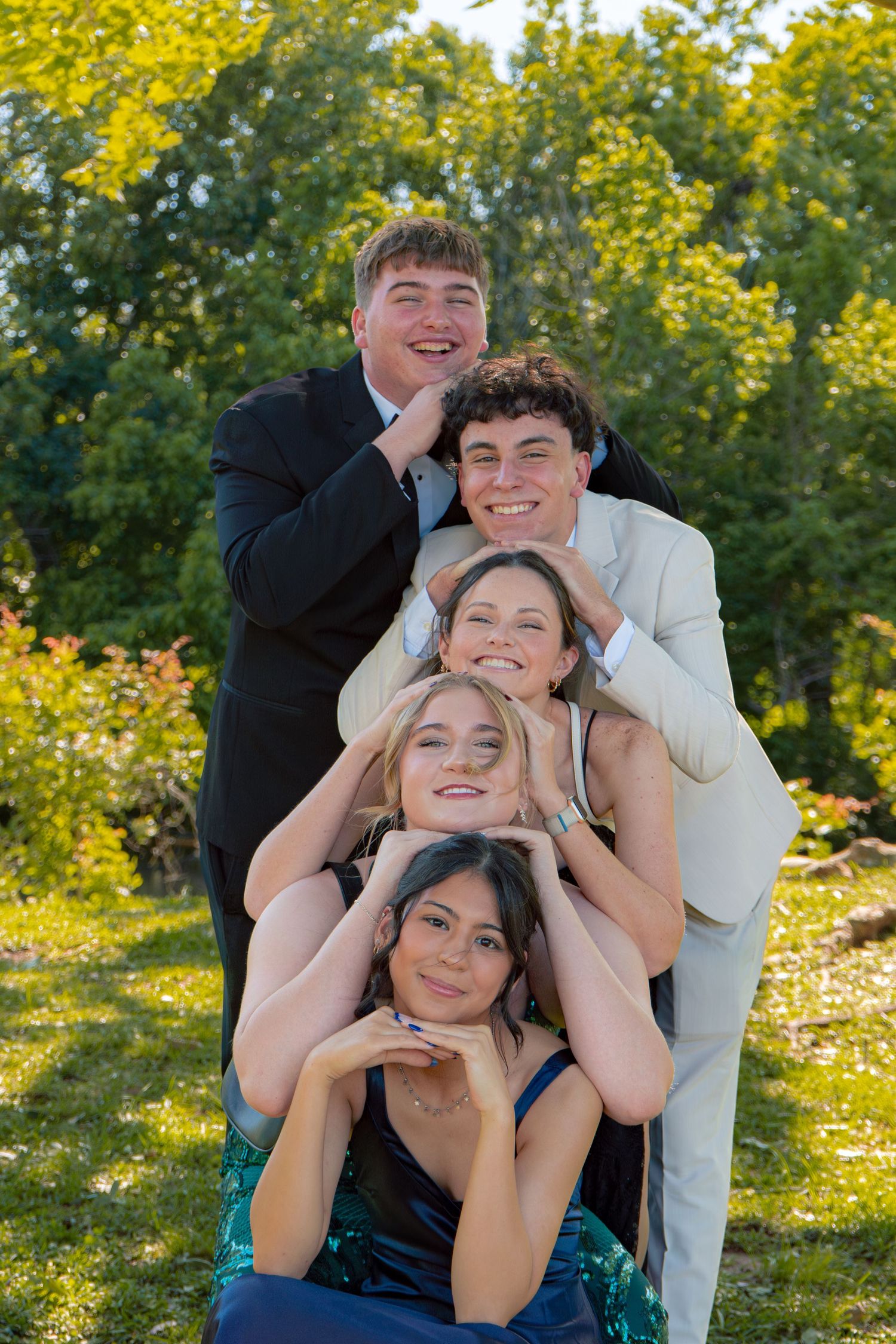 Five prom-dressed teenagers pose in a fun stacked portrait formation outdoors in a sunny green park.