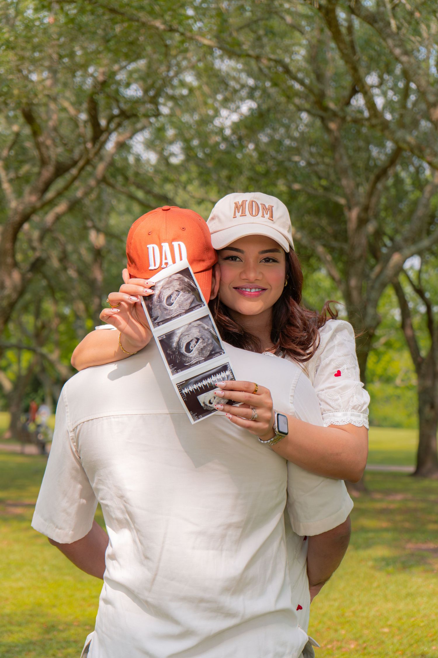 Pregnant couple hugging outdoors, woman wearing MOM cap holds ultrasound photo while man wears DAD cap.