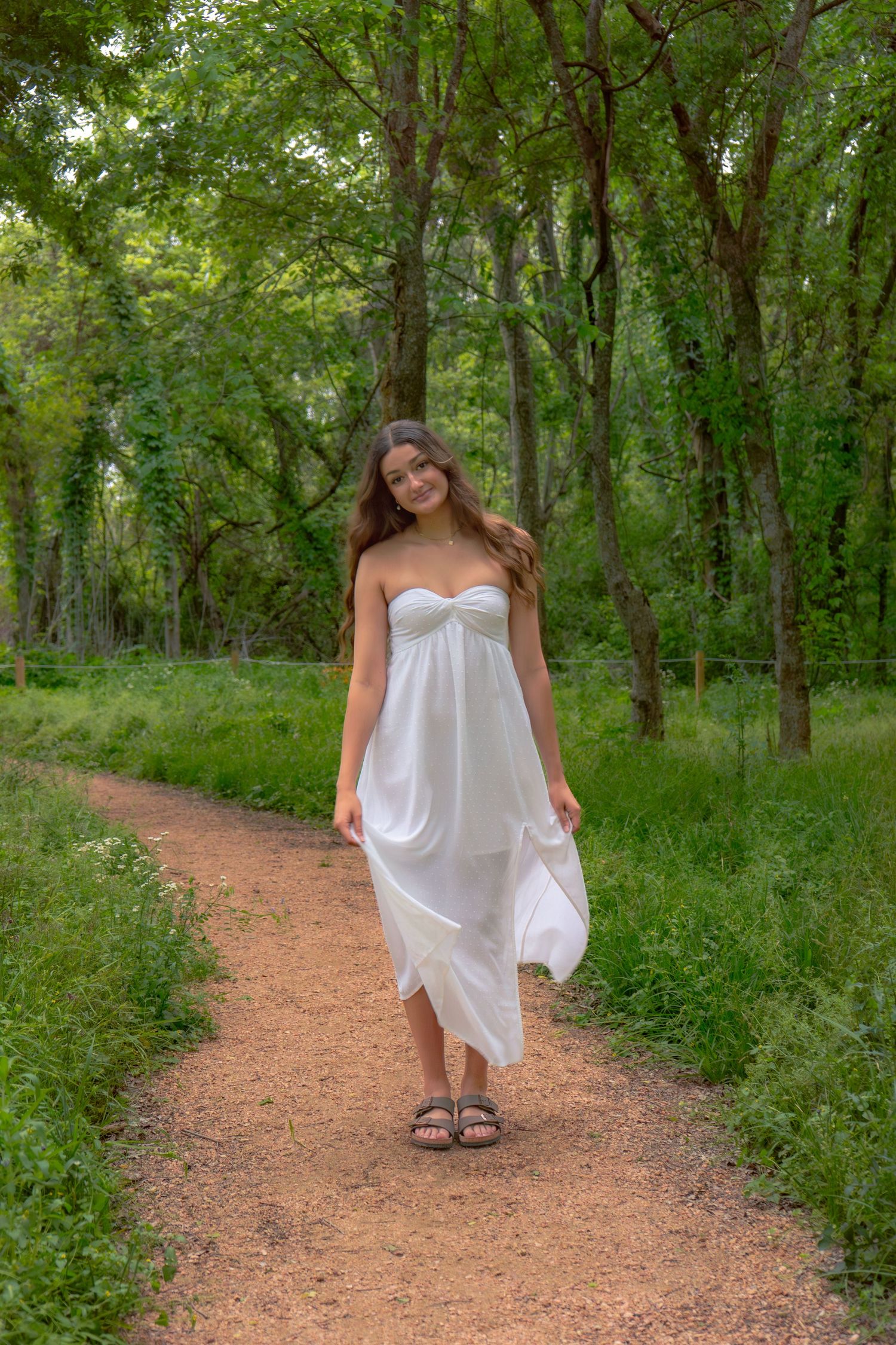 Woman in flowing white strapless dress walking on a dirt path through green woods.