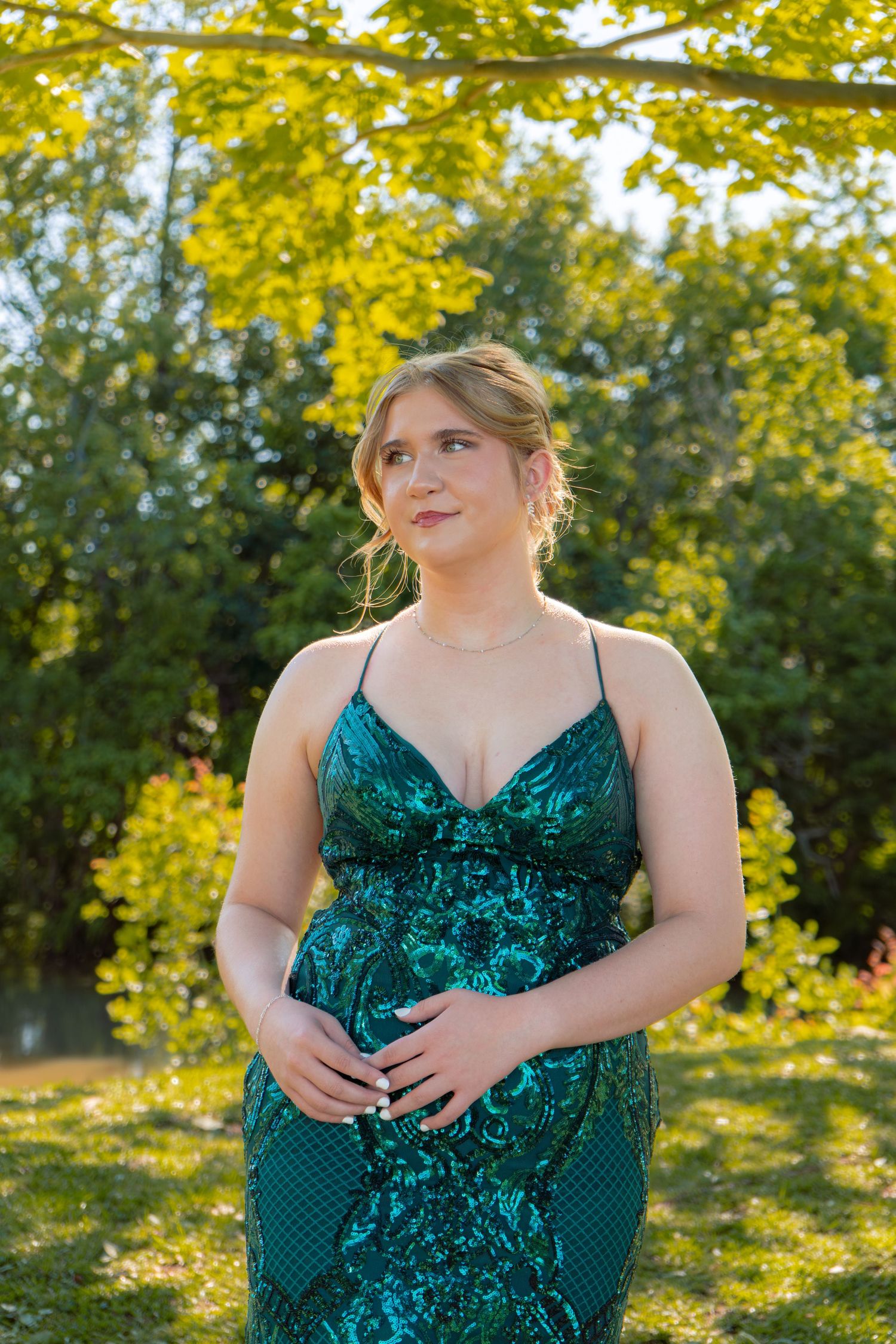 Blonde woman in emerald green sequin spaghetti strap formal gown posing under yellow-green trees outdoors.