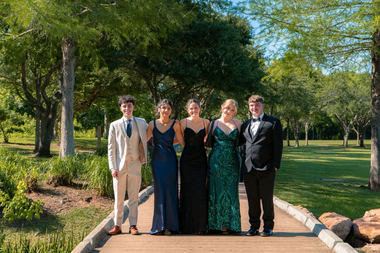 Five teenagers in formal prom attire pose together on a pathway surrounded by lush green trees and gardens.