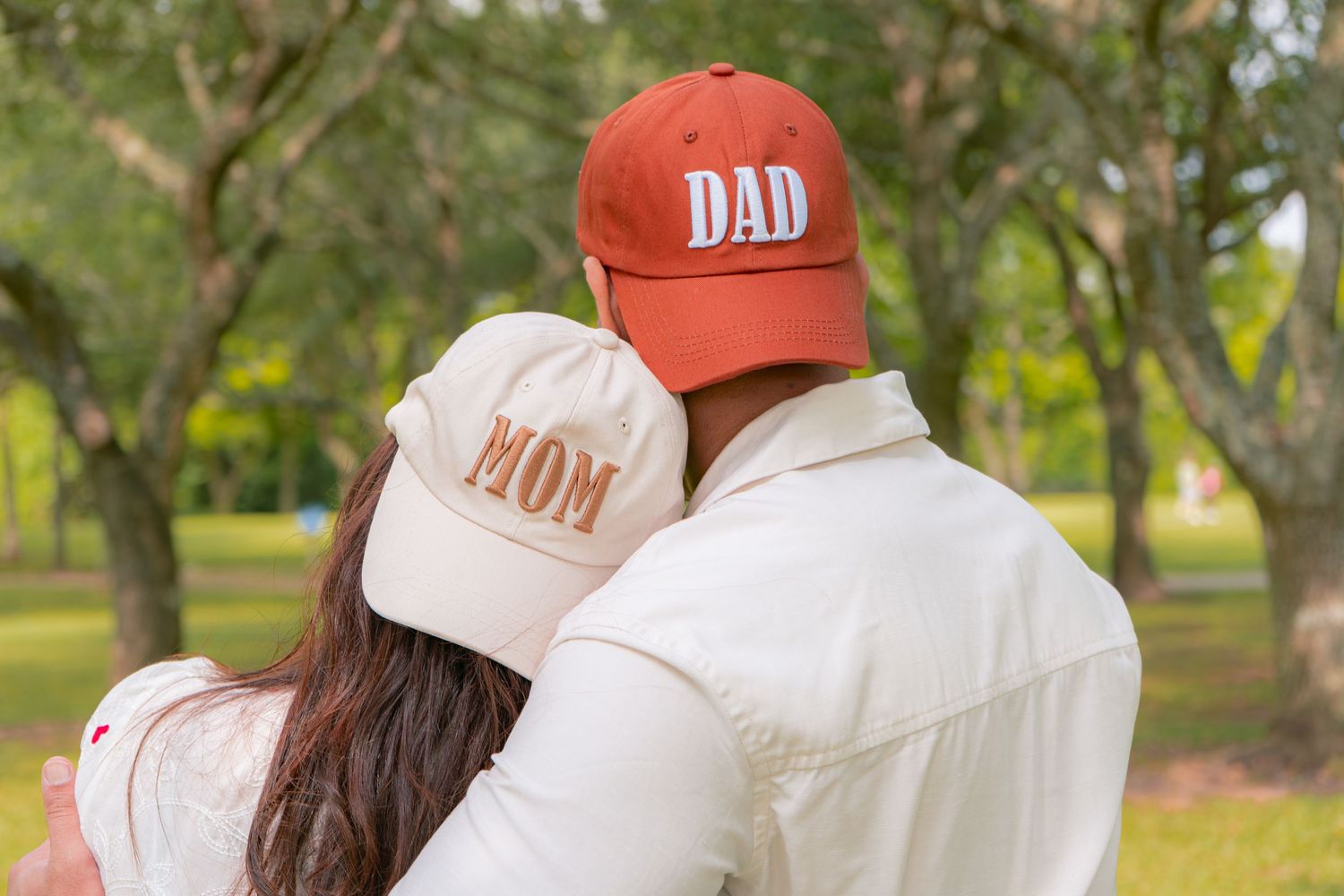 Couple embracing outdoors wearing matching Mom and Dad baseball caps in park setting.