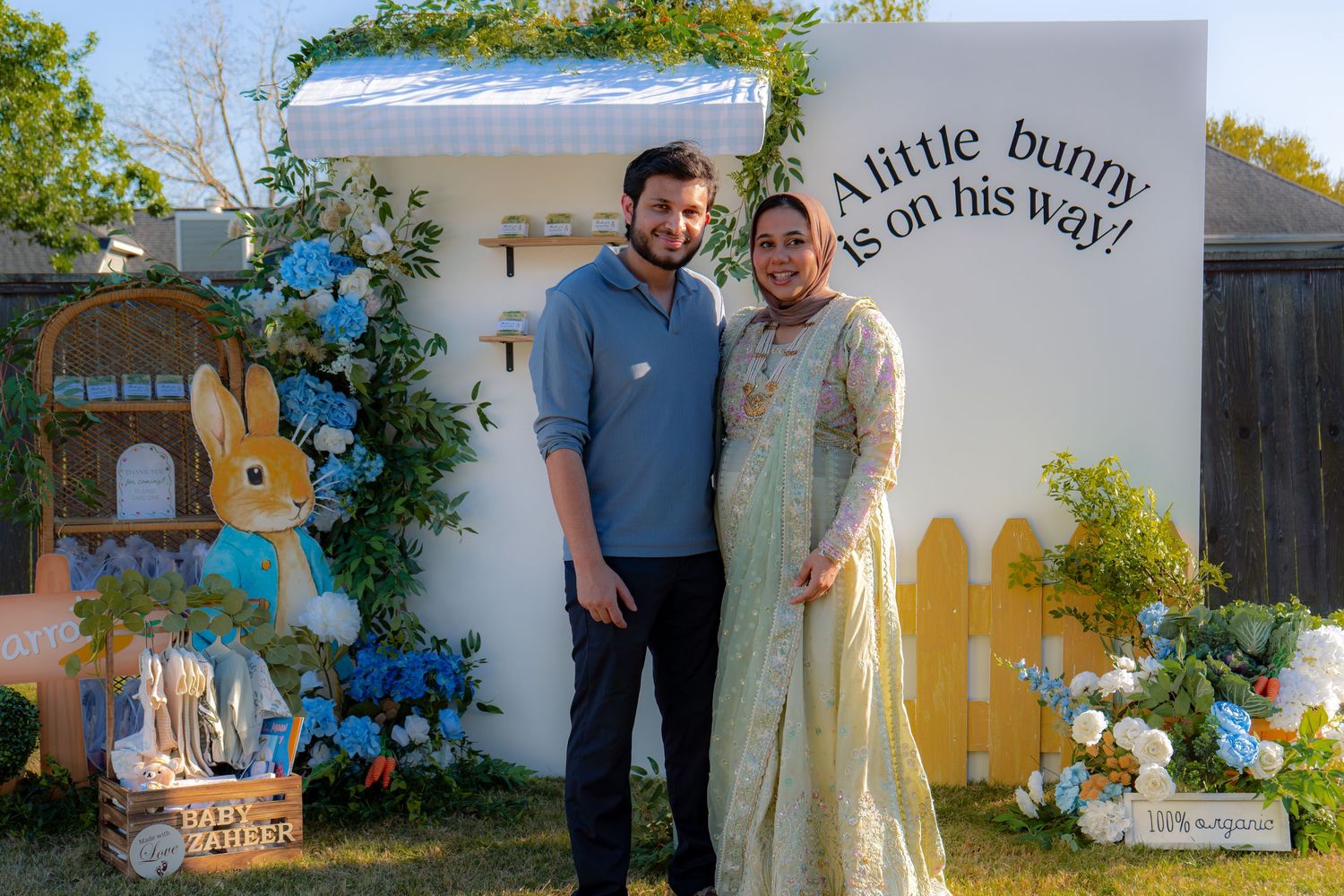 Couple posing at Peter Rabbit themed baby shower backdrop reading A little bunny is on his way.