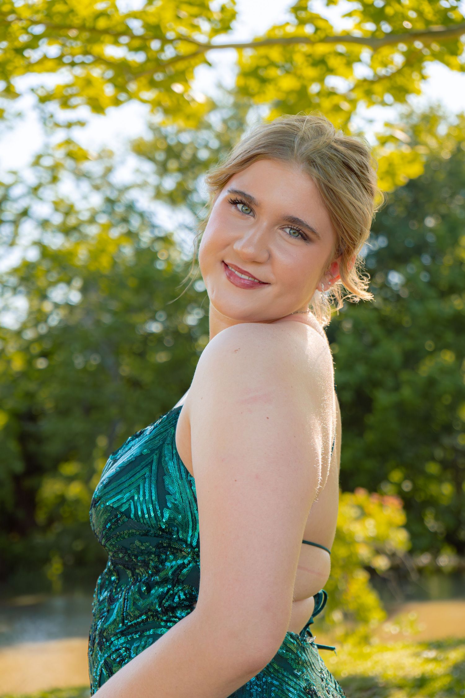 Young blonde woman in a green sequined prom dress glances over her shoulder outdoors in golden sunlight.