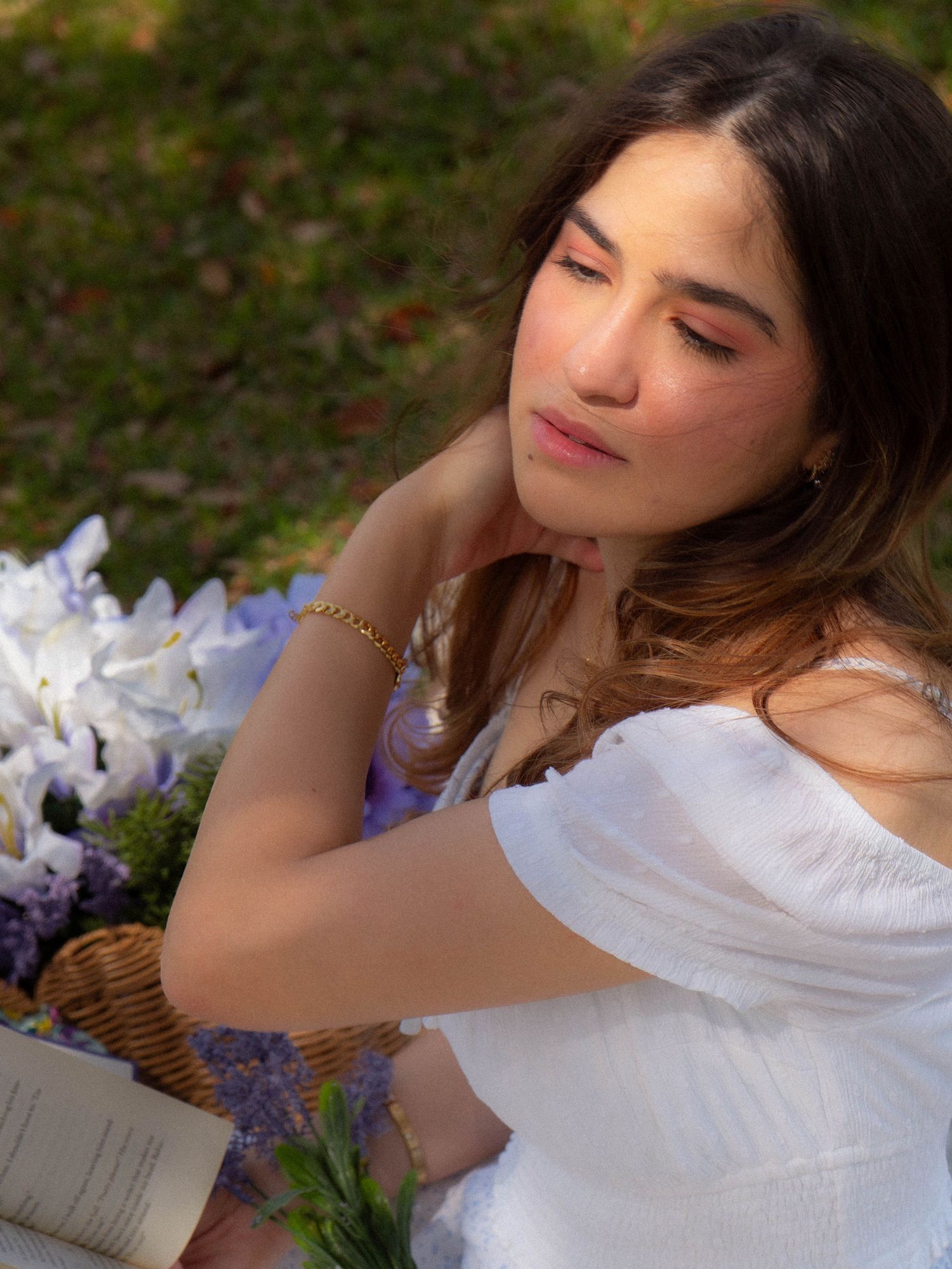 Young woman in white off-shoulder dress smelling purple flowers from wicker basket during outdoor picnic.