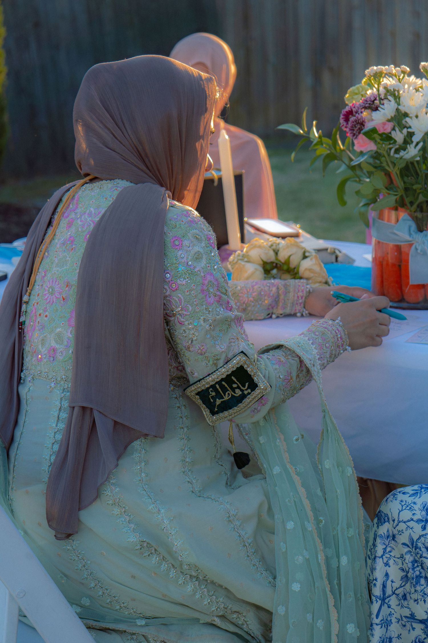 Woman in embroidered mint green salwar kameez and taupe hijab seated at an outdoor table.