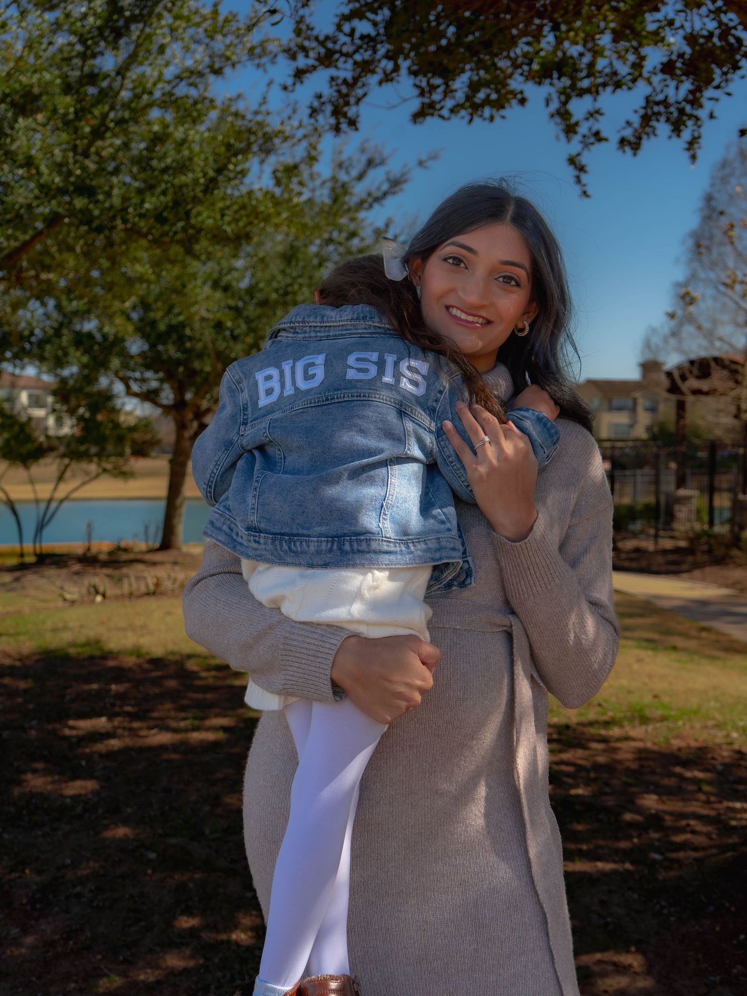 Smiling pregnant woman holding toddler in Big Sis denim jacket outdoors in park.