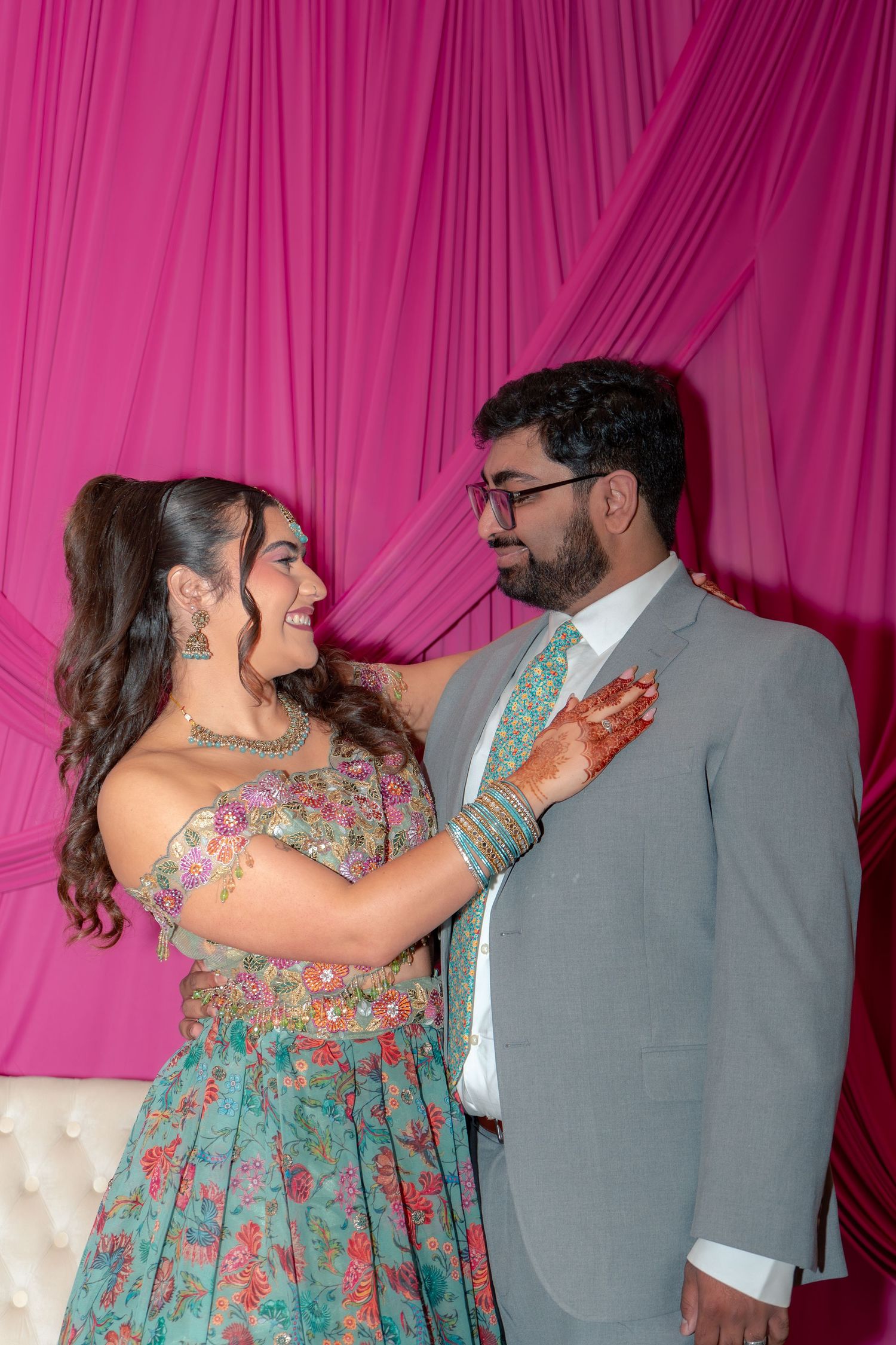 Smiling South Asian bride in teal lehenga gazing at groom in grey suit with green tie against pink draped backdrop.