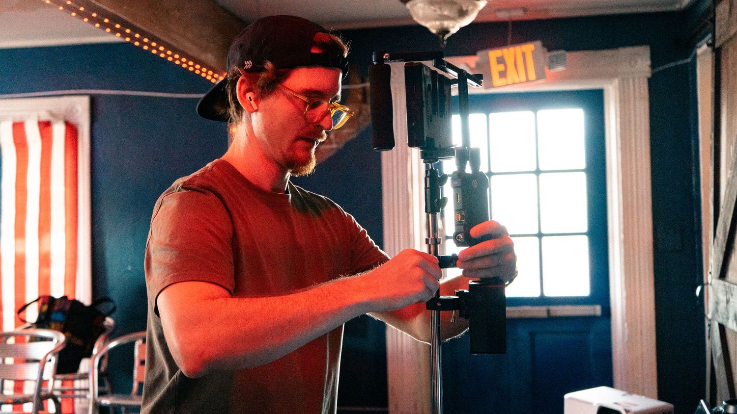 A young man in a red shirt and cap adjusts film equipment indoors during a video production shoot.