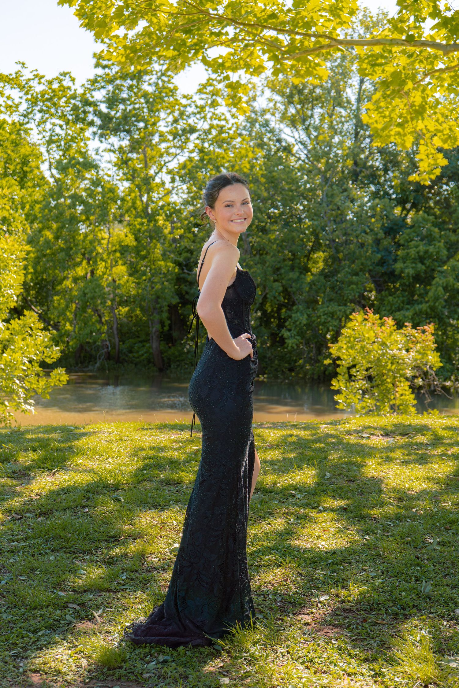 Woman in stunning backless black sequin formal gown posing sideways in lush green park at golden hour.