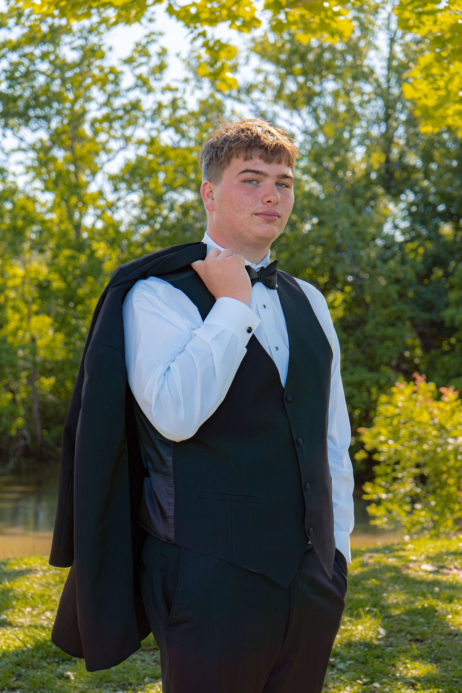 Young man in tuxedo with bow tie drapes jacket over shoulder outdoors among green trees.