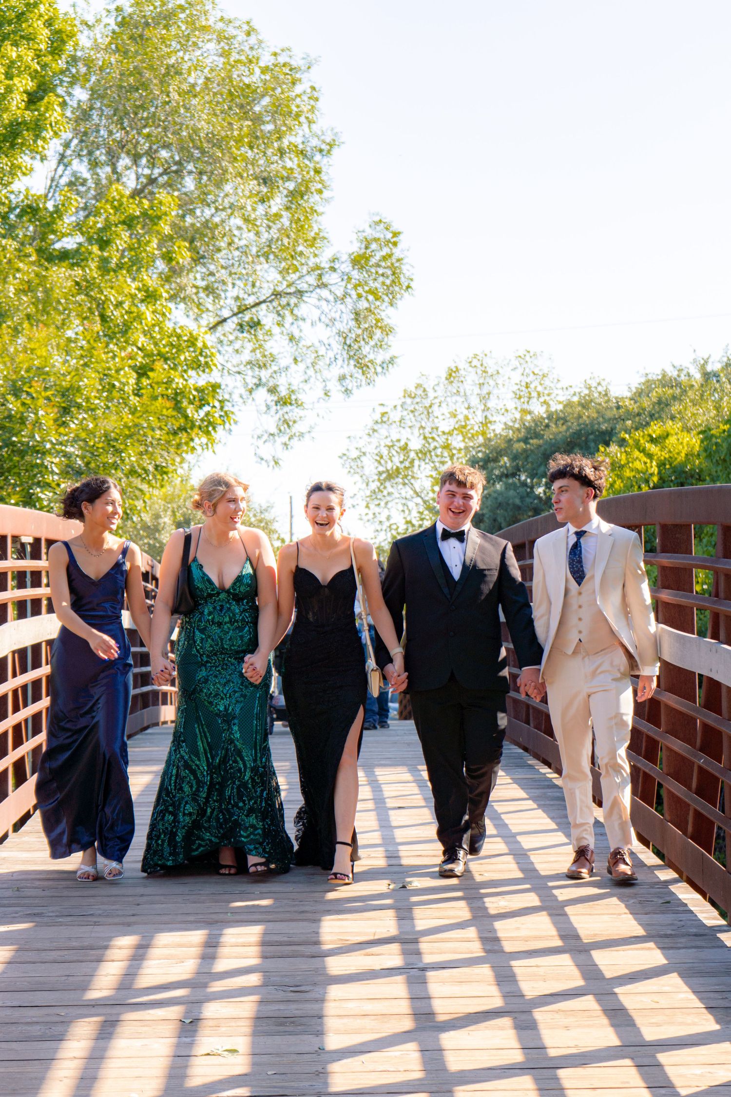 Five prom-dressed teens walk hand-in-hand across a sunny wooden bridge surrounded by green trees.