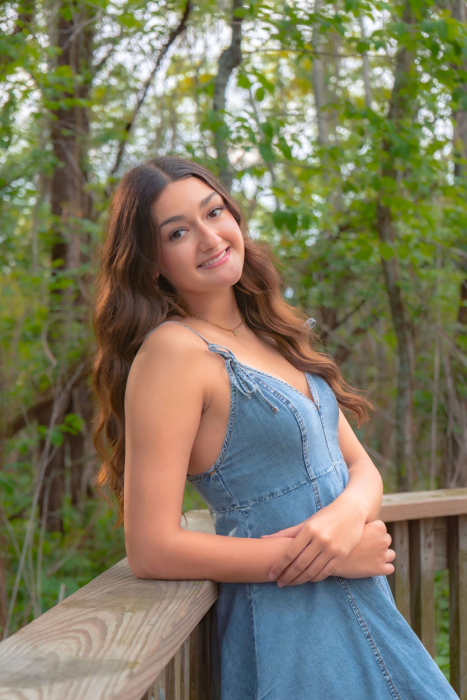 Young woman in denim dress leaning on wooden railing, smiling outdoors with lush green forest background.