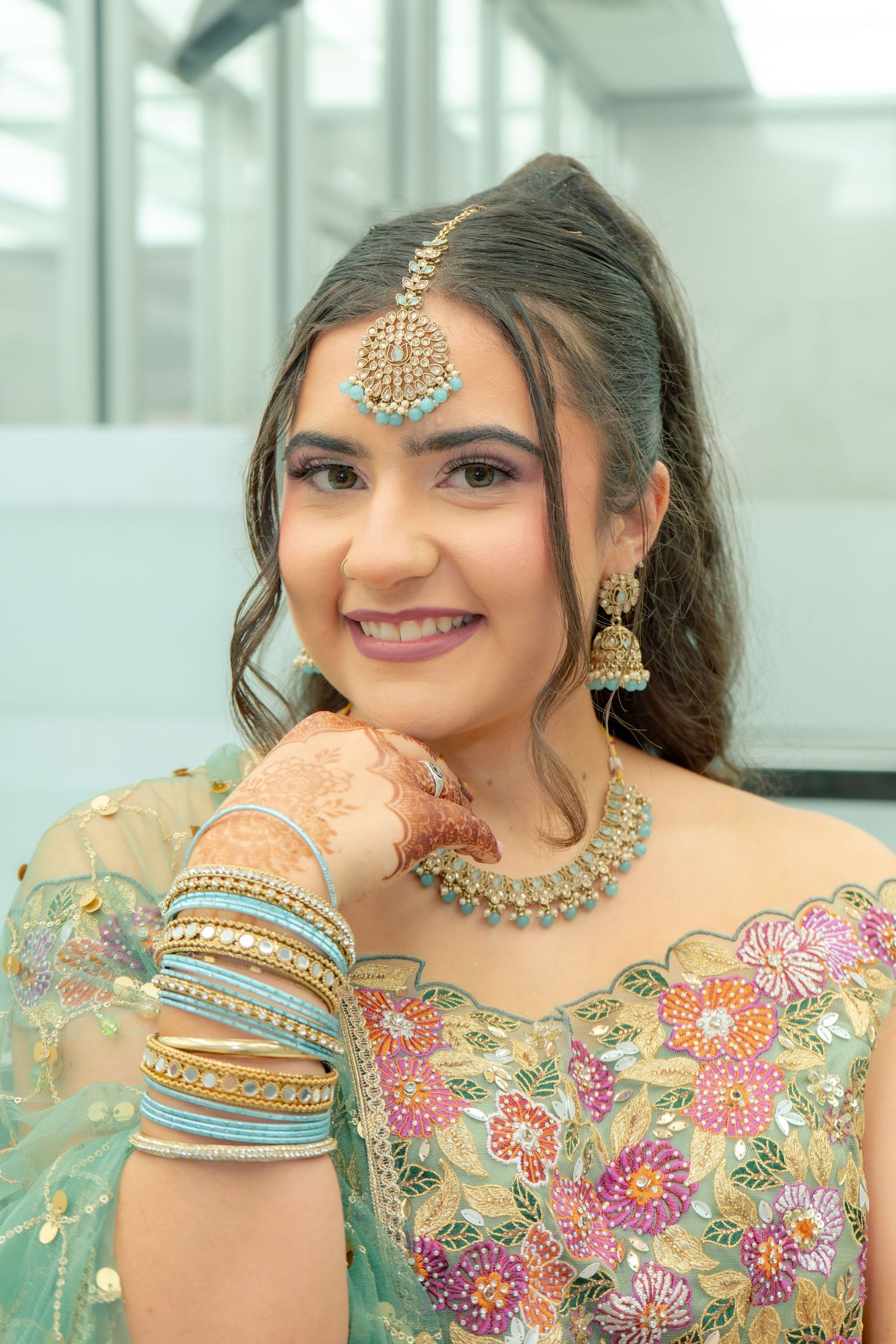 Young South Asian bride in teal floral lehenga with gold maang tikka, jhumka earrings, bangles, and mehndi smiling at camera.