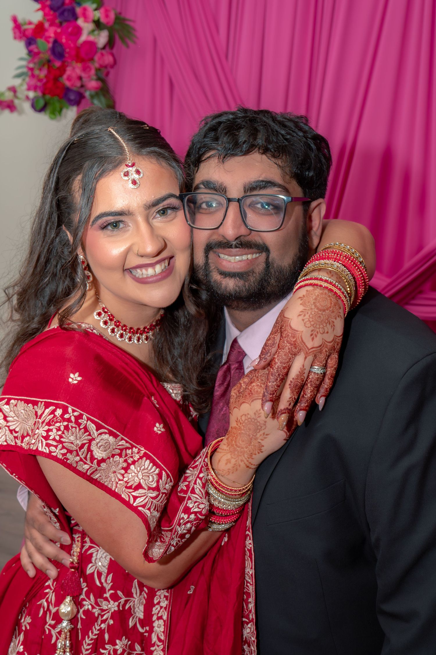 Smiling Indian couple in traditional wedding attire posing together against pink floral backdrop.