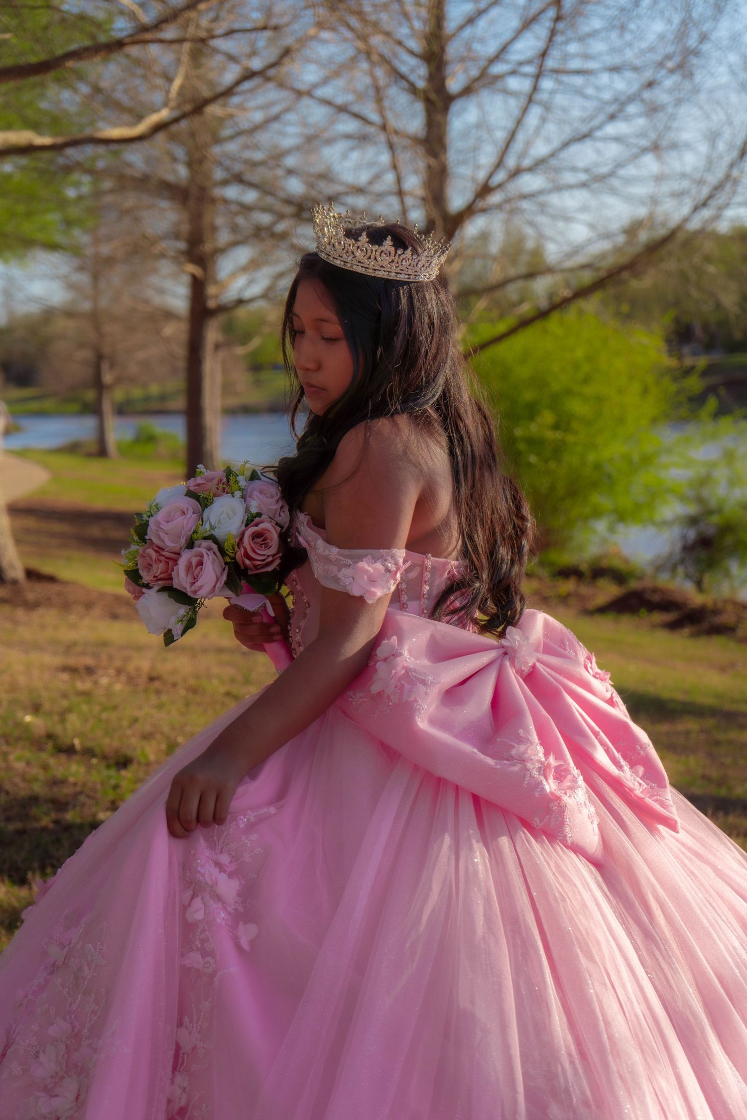 Young girl in pink quinceañera ball gown with crown holds bouquet in park setting.