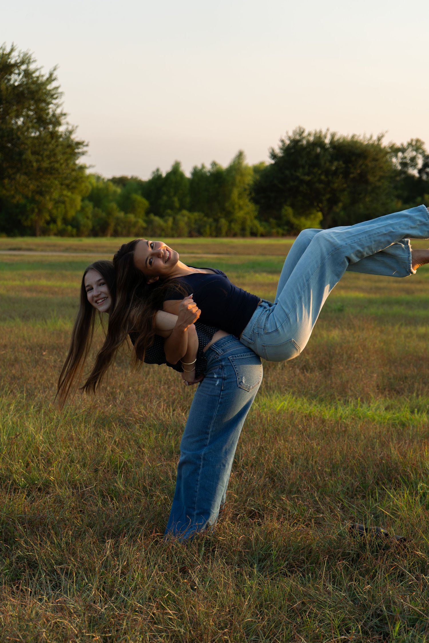 Two laughing young women in jeans play outdoors at sunset, one lifting the other in a grassy field.