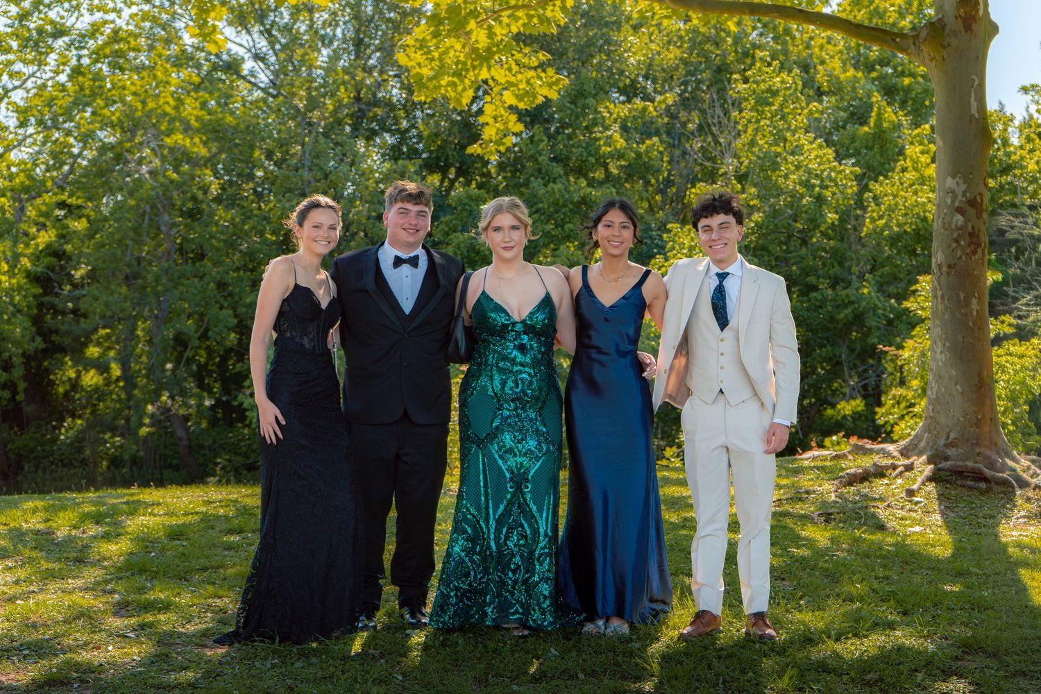 Five teenagers in formal prom attire pose together outdoors on green grass with trees in the background.