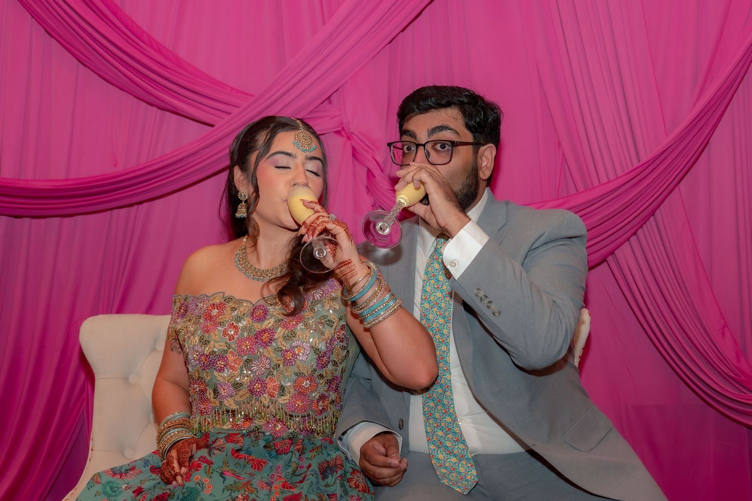 Couple in festive attire eating snacks in front of pink draped backdrop at Indian celebration.