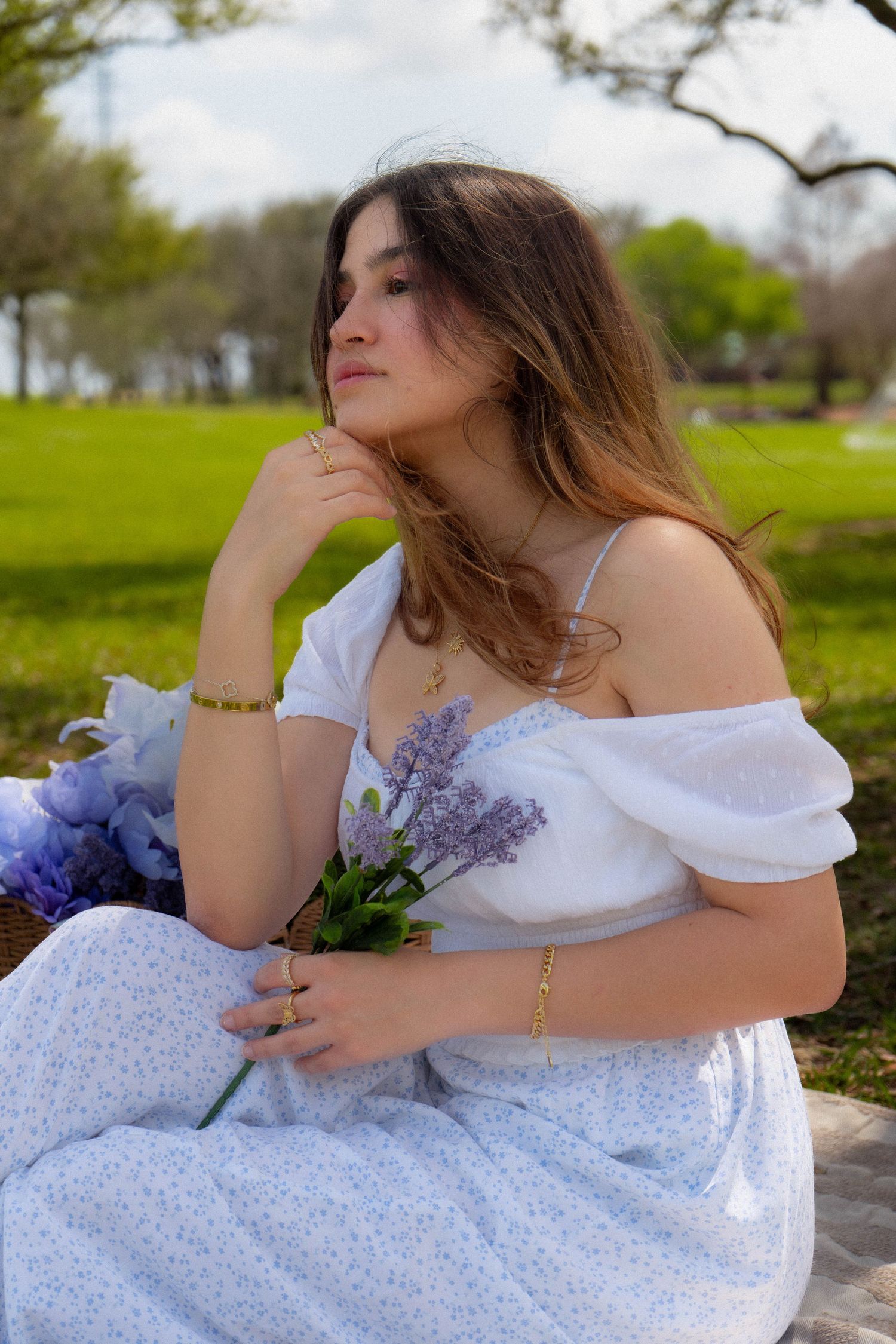 Thoughtful young woman in white off-shoulder dress holding lavender flowers while sitting in green park.