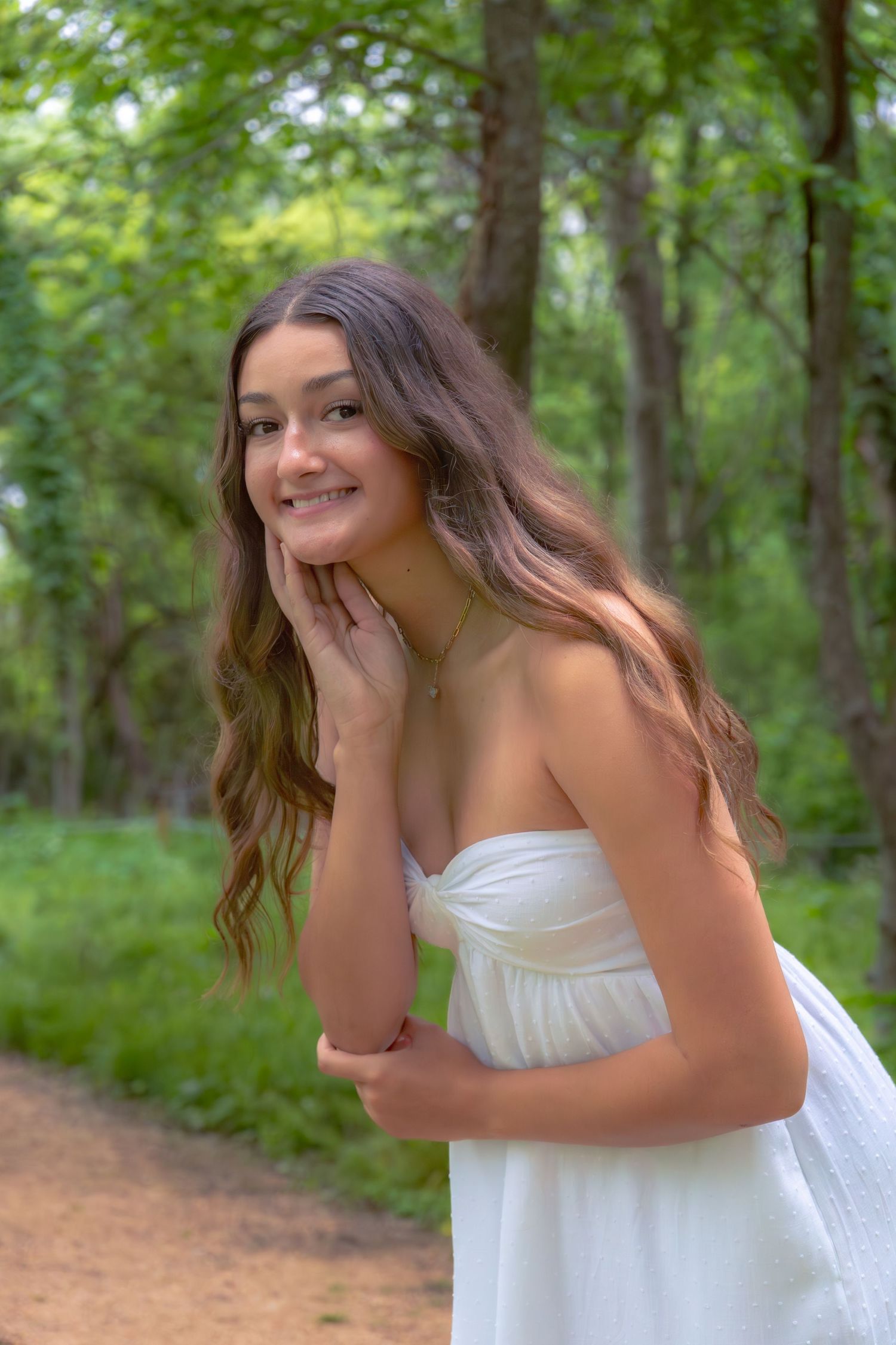 Young woman in white strapless dress smiling in a lush green forest path.