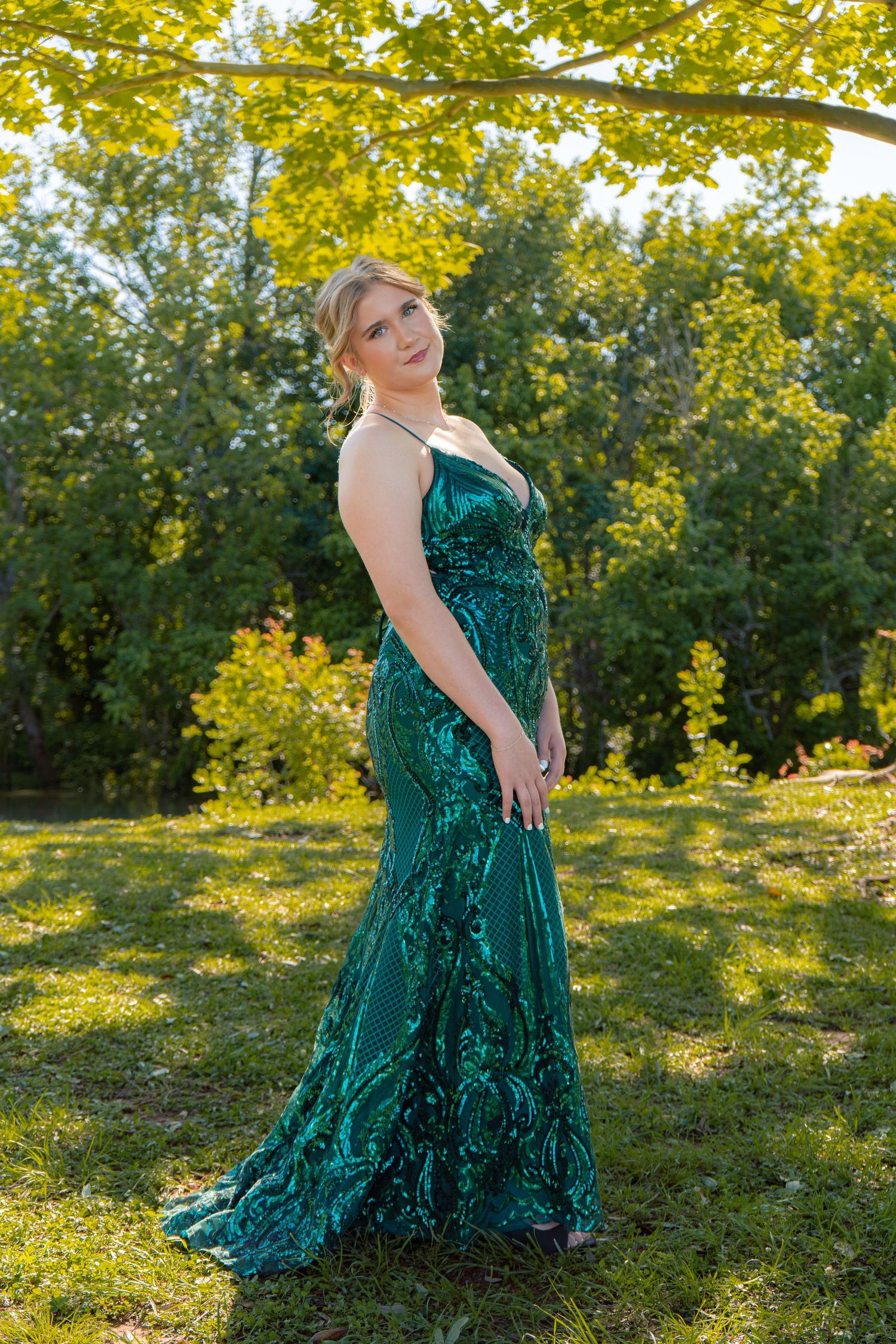 Woman in elegant emerald green sequin mermaid gown posing outdoors in lush green garden setting.