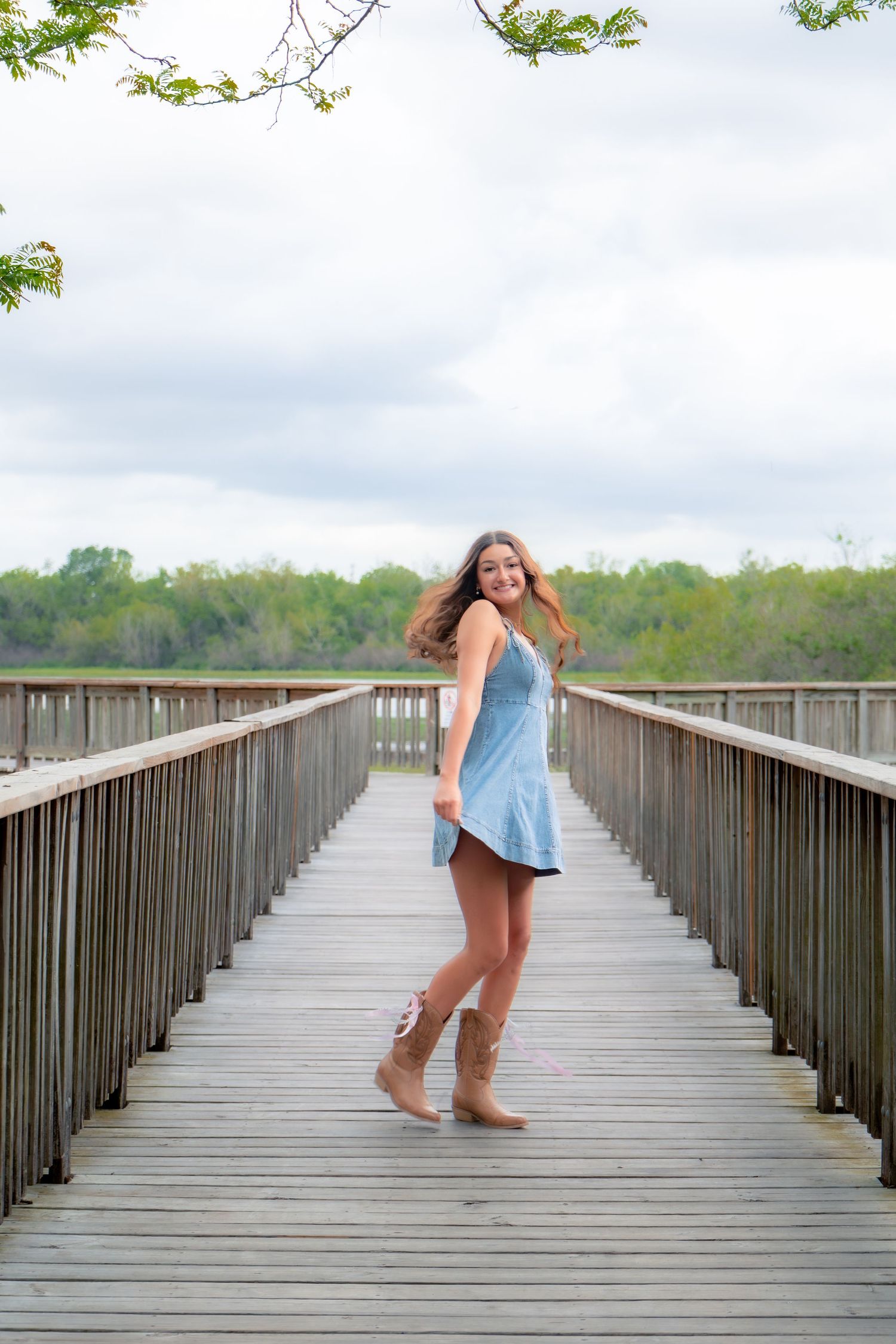 Smiling young woman in blue denim dress and tan cowboy boots twirling on a wooden bridge walkway.