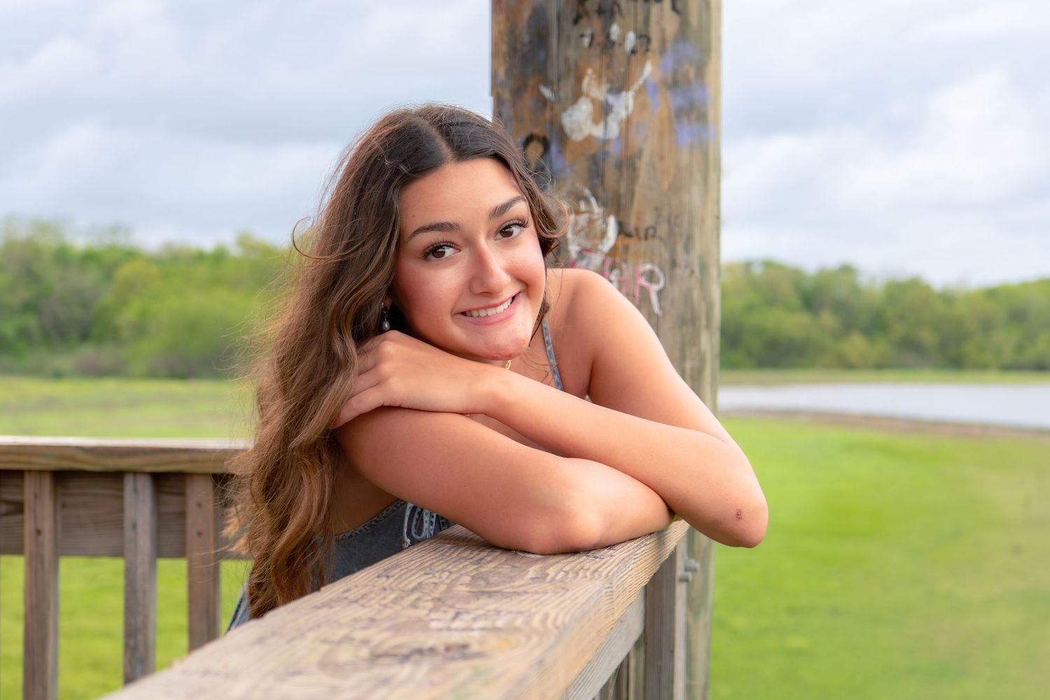 Young woman with long wavy hair smiling while leaning on a wooden railing outdoors near a green field.