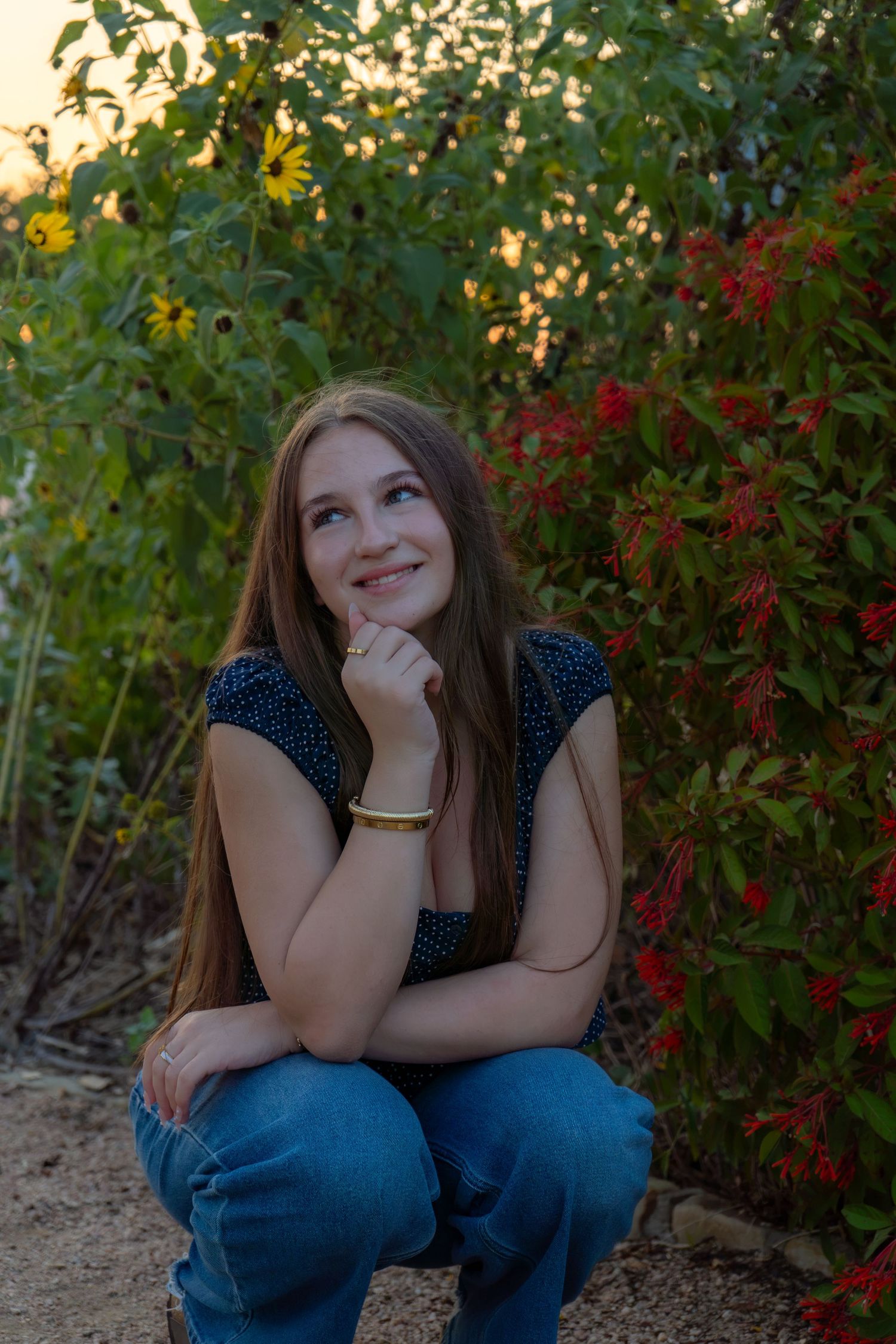 Young woman crouching in garden with red and yellow flowers, smiling thoughtfully with hand on chin at sunset.